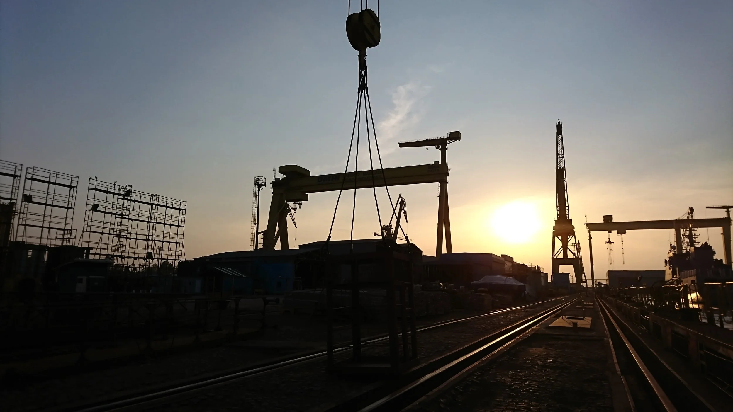 Industrial shipyard at sunset with cranes, docked ships, and railway tracks.