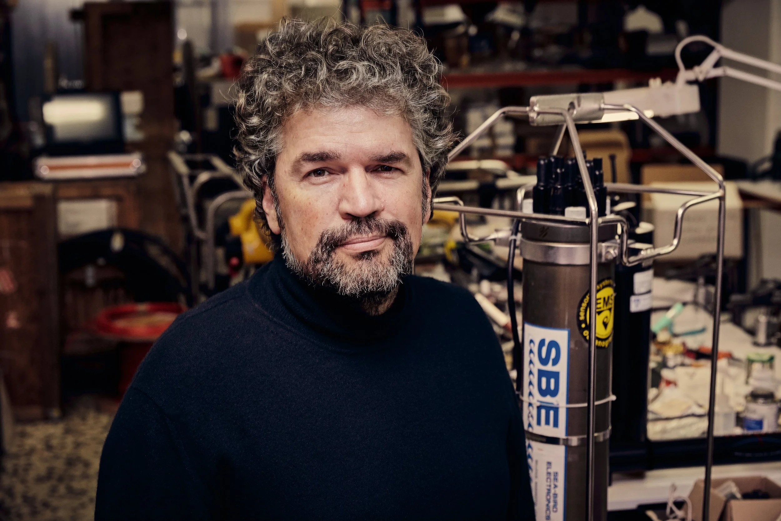 A man with curly gray hair and a beard wearing a black turtleneck, standing in a workshop or laboratory environment, with various tools and equipment in the background.