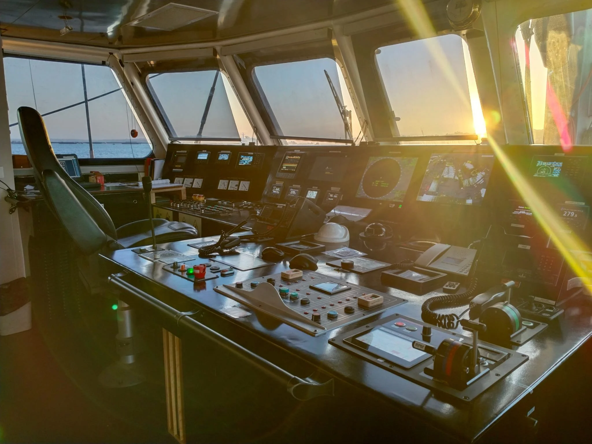 The image shows a ship's bridge with various navigational and communication instruments, a large window view of water and a setting sun, and a captain's chair.