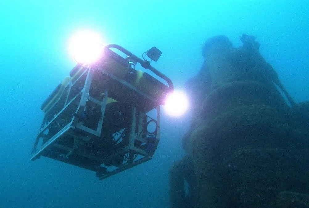 Underwater scene with a remote-operated underwater drone illuminated by lights, next to a submarine hull.