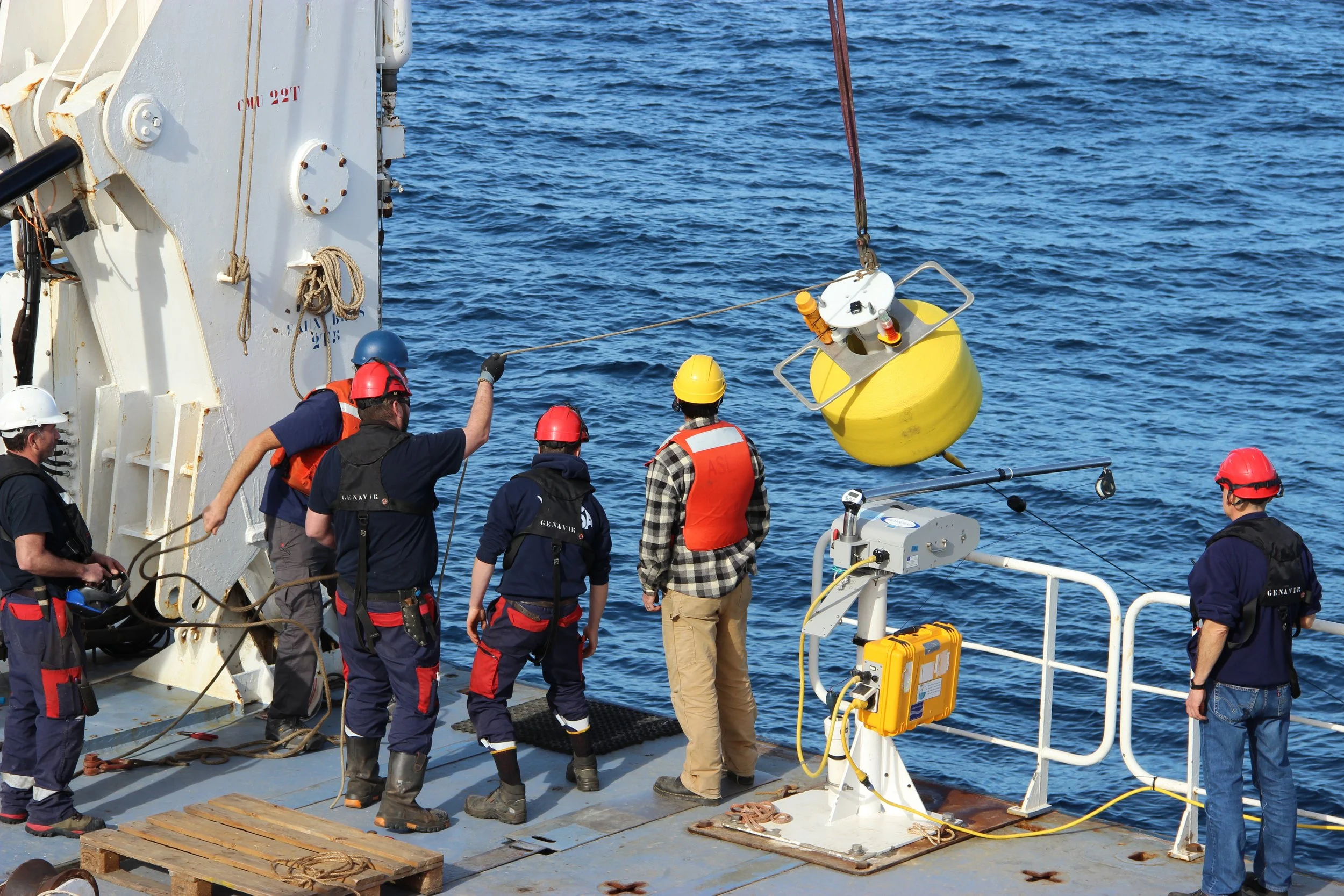 Ship crew members in safety gear lowering a yellow buoy into the water from a ship.