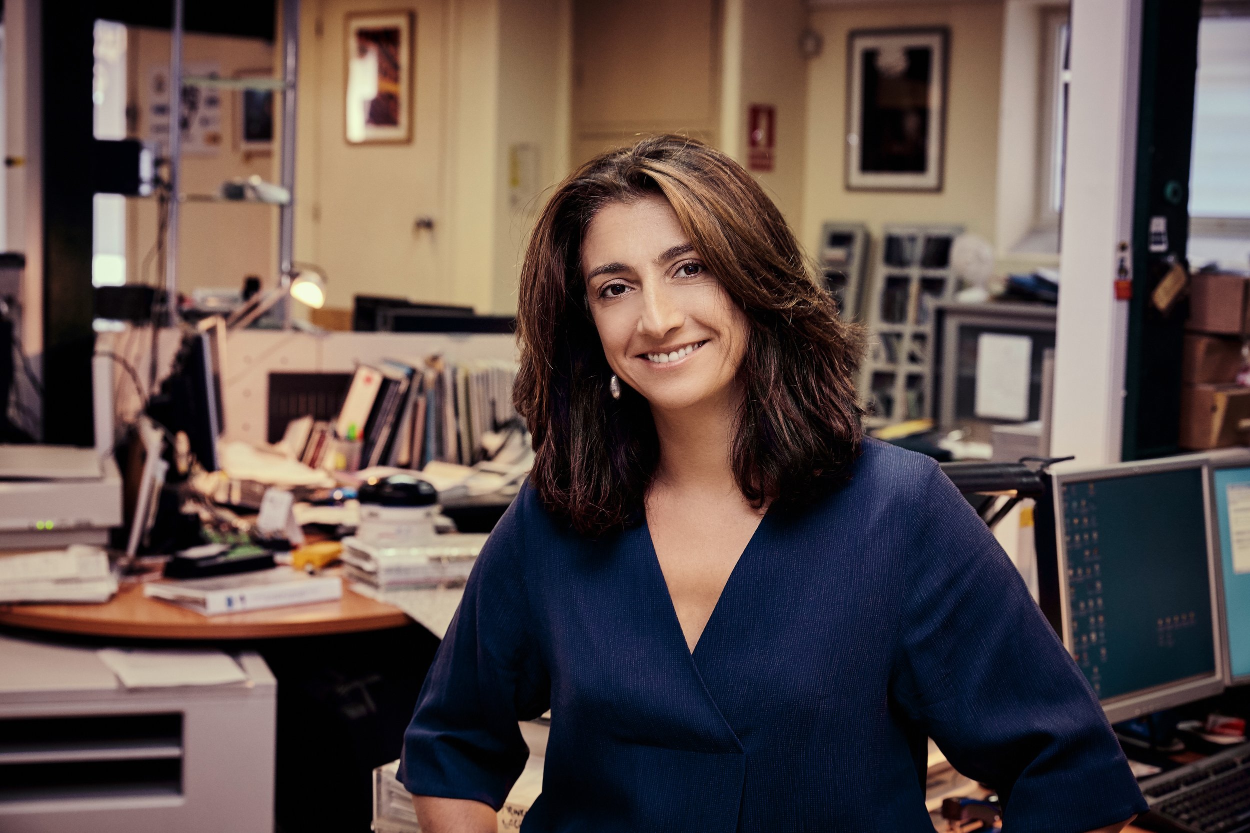 A woman with shoulder-length brown hair smiling in an office space filled with desks, computers, and stacks of papers.