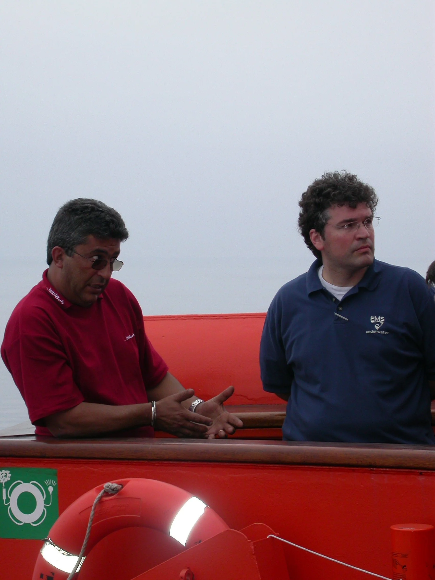 Two men standing on a boat with an orange exterior, one wearing a red shirt and sunglasses, the other wearing a navy blue shirt with a logo, on a cloudy day.