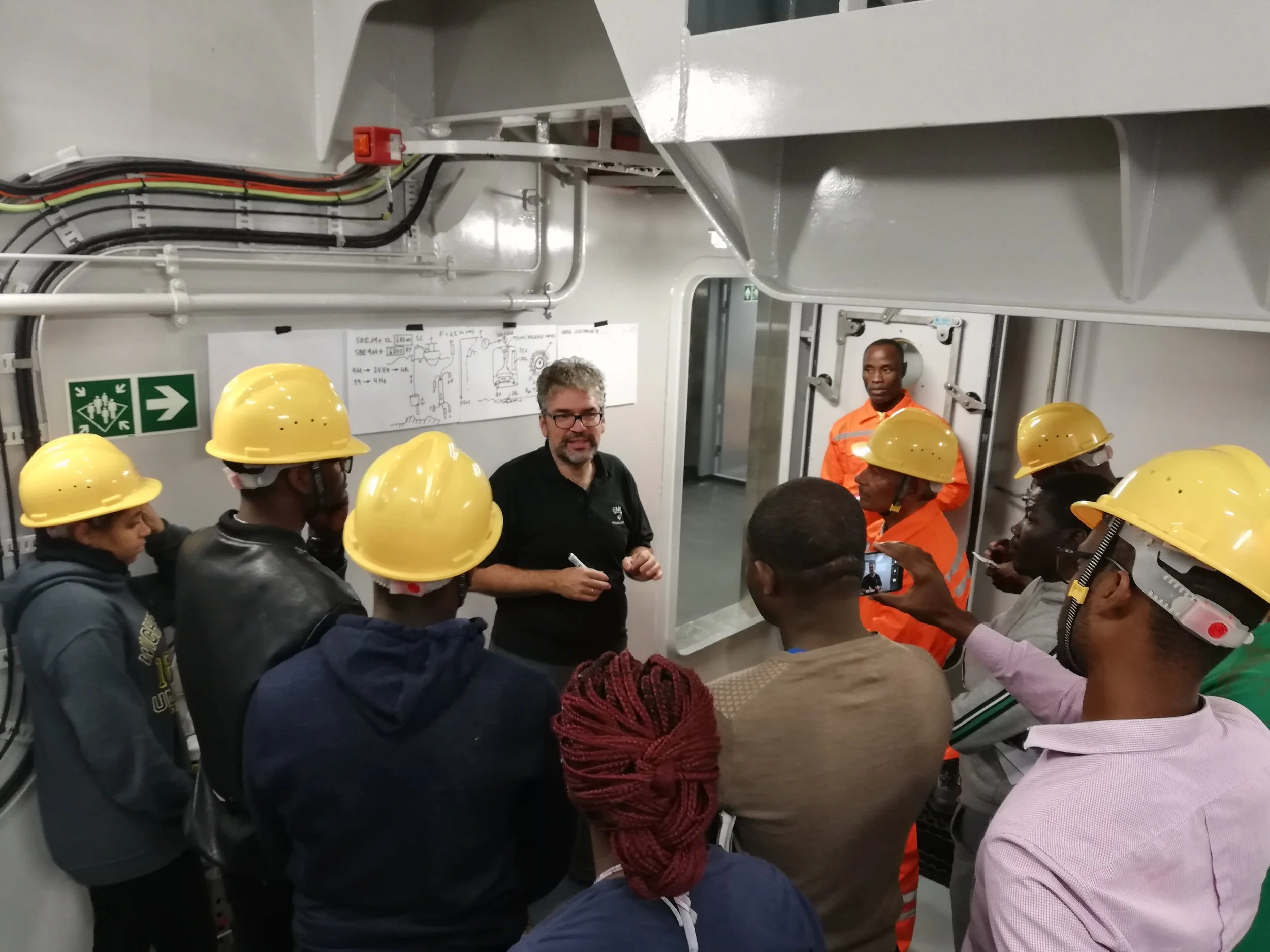 A group of people, wearing yellow safety helmets, are gathered around a man who appears to be explaining something. The group includes individuals in casual clothing and safety uniforms, with a woman taking a photo. The setting looks like an industri