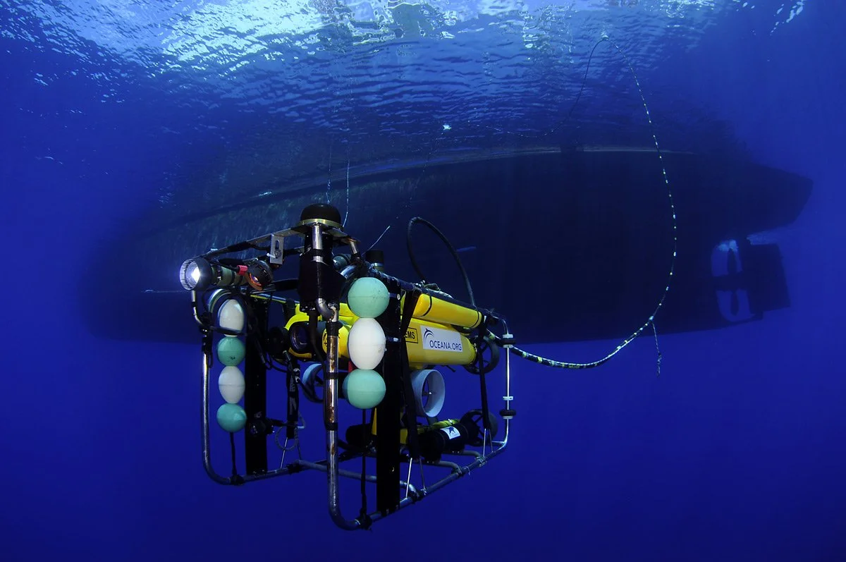 A submarine underwater with a remotely operated vehicle (ROV) in the foreground, carrying equipment including floats and cables, with a large shadow of the submarine visible beneath the water surface.