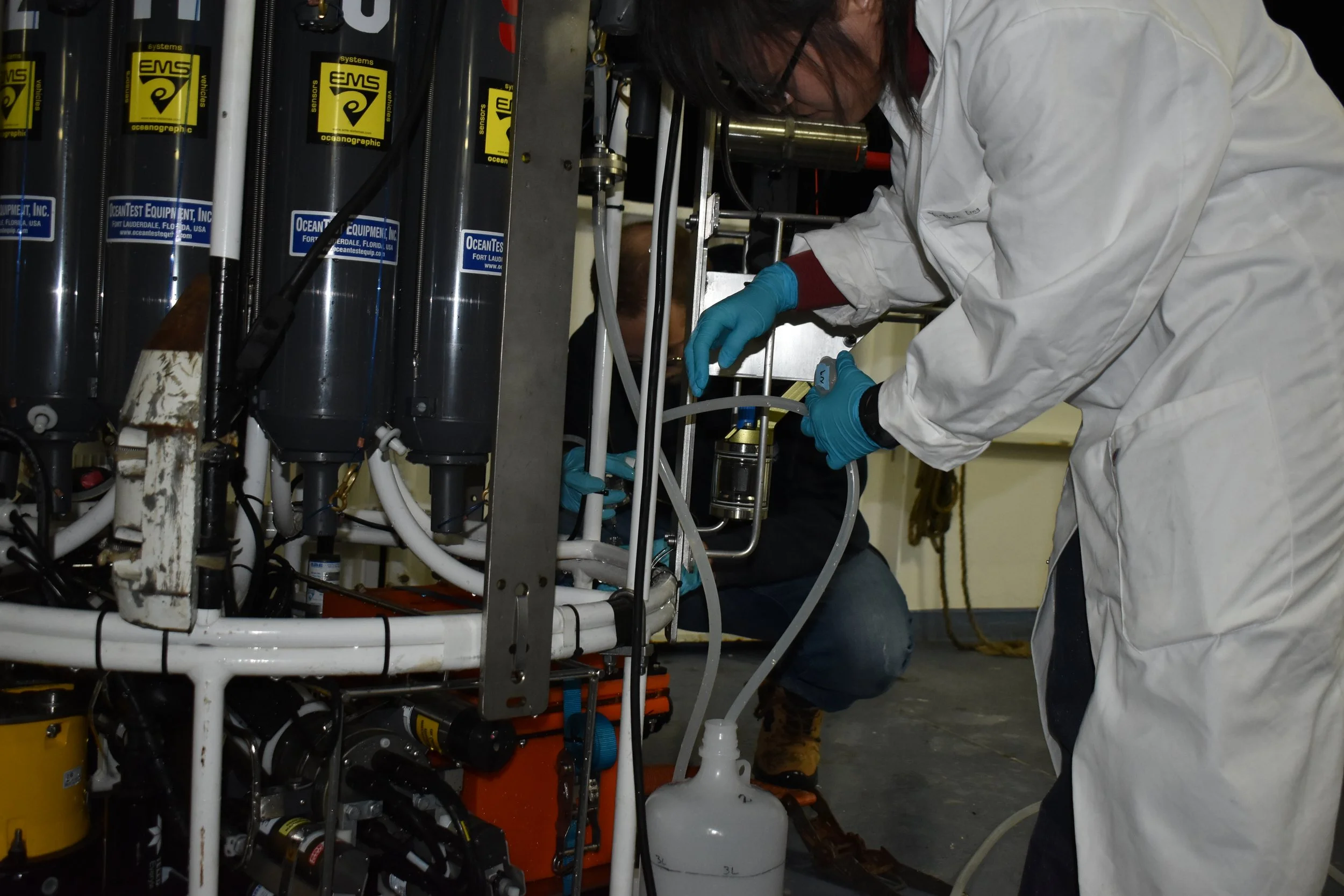Scientists in protective white coats working with laboratory equipment, including large stainless steel cylinders and tubes, in a laboratory setting.