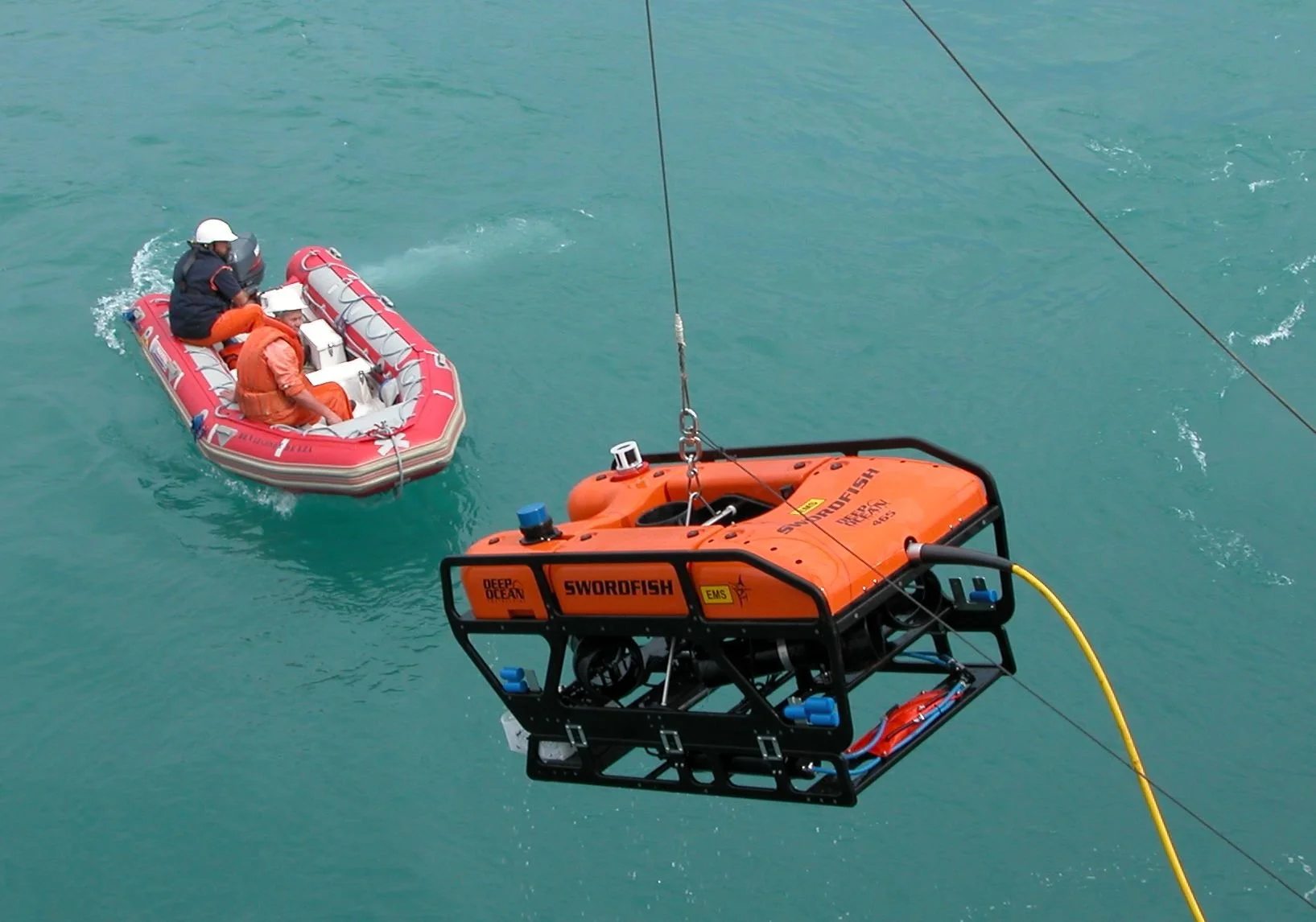 Two people in orange and black gear on an inflatable boat near an orange submerged vehicle with the label 'Swordfish' below water surface