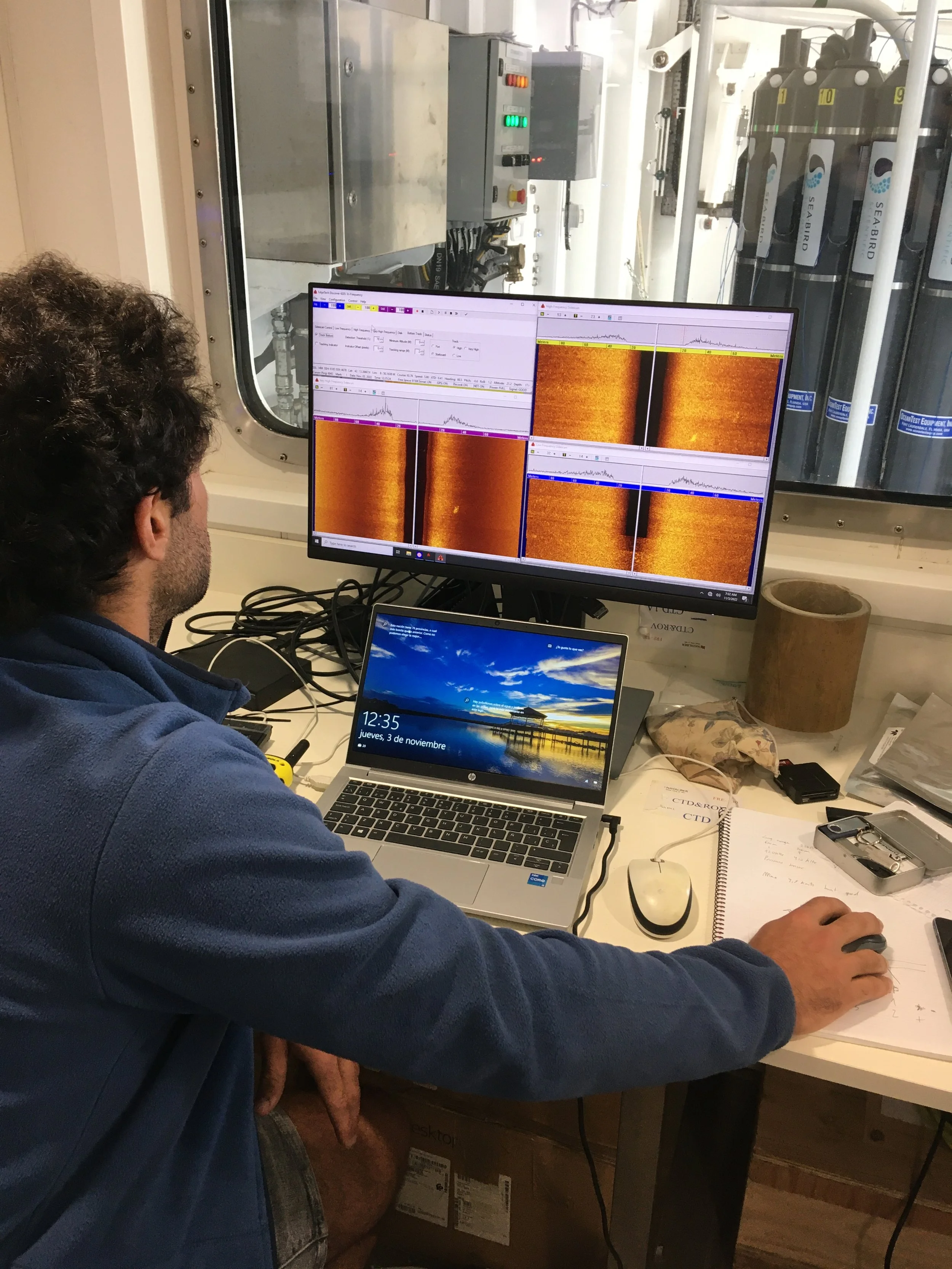 A person working at a desk with a laptop, a mouse, and a notepad, viewing a computer monitor displaying scientific data, with a window showing equipment outside.