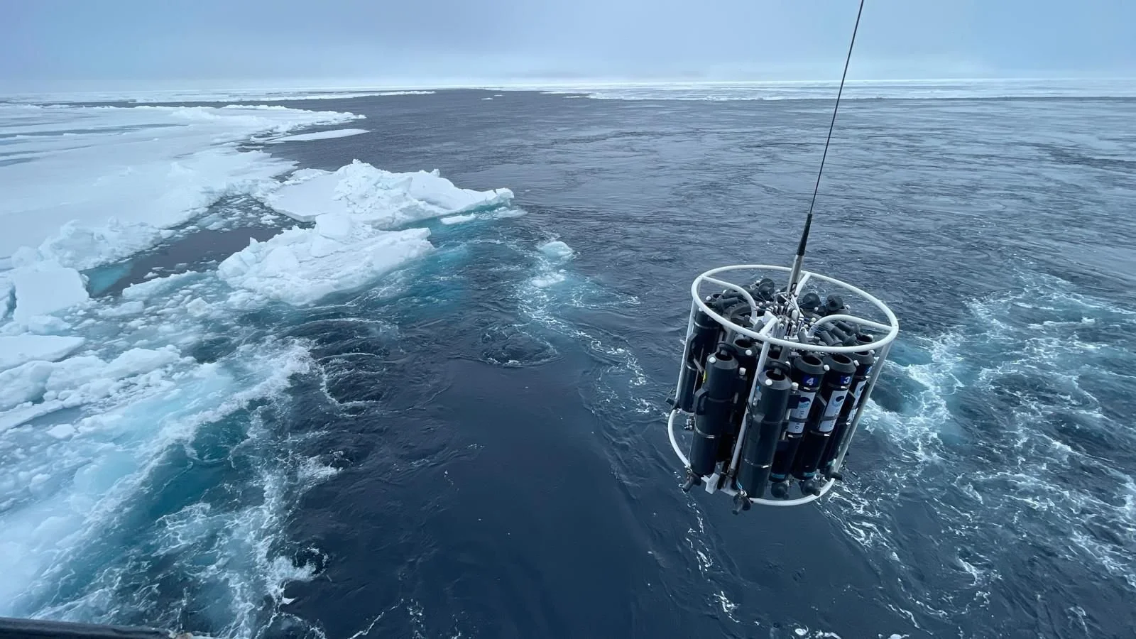 Research equipment suspended over icy Arctic waters with floating ice chunks and snow-covered ice in the background.