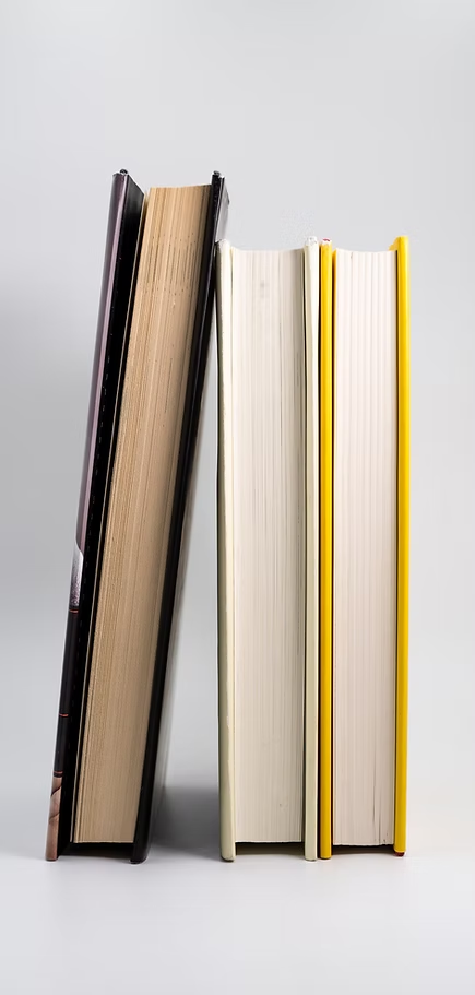 Four books standing upright on a white surface against a white background, including a magazine, a science textbook with a black cover, a white hardcover book, and a yellow-edged paperback.