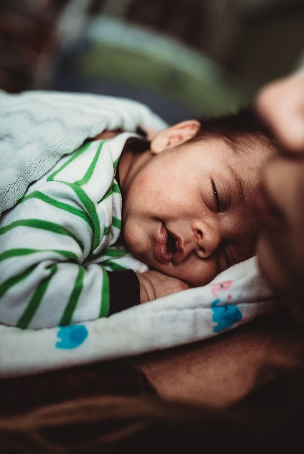 A newborn baby sleeping on an adult's chest, wrapped in a blanket, wearing a striped onesie with green and white lines.