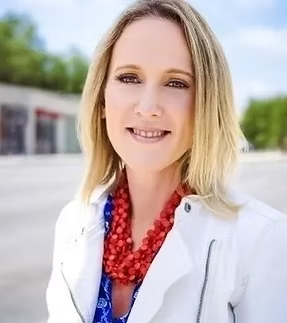 A woman with blonde hair wearing a white jacket and red necklace, smiling outdoors with trees and a building in the background.
