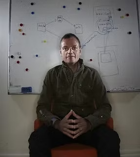 A man sitting in front of a whiteboard with various diagrams and notes drawn in black marker.