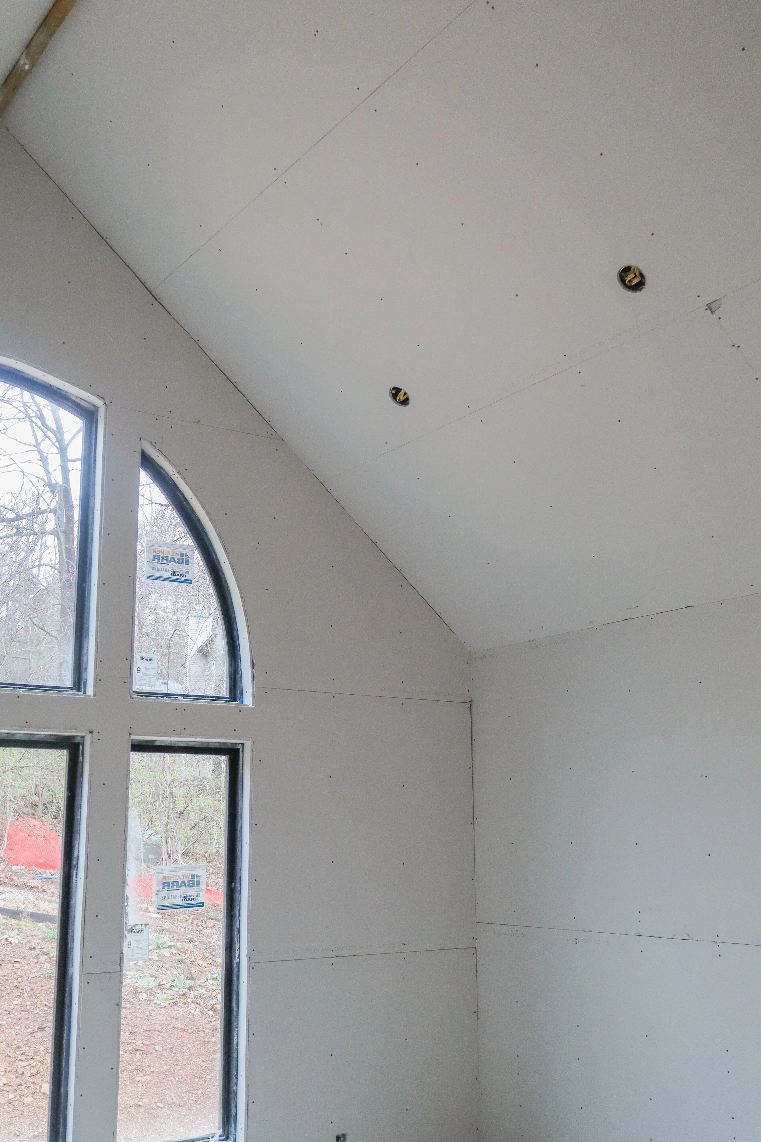 Interior under construction with drywall being installed on walls and ceiling, with electrical wiring visible through open ceiling fixtures, and large arched windows revealing an outdoor wooded area.