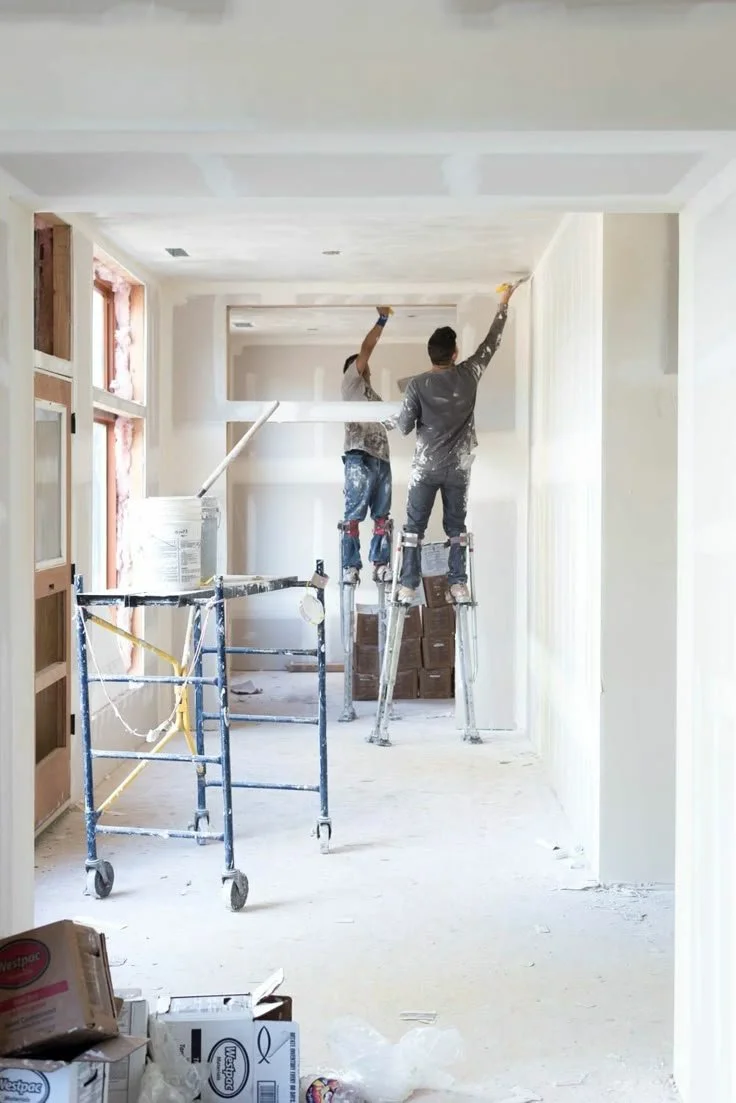 Two workers are painting a ceiling in a room under construction, standing on ladders and scaffolding.