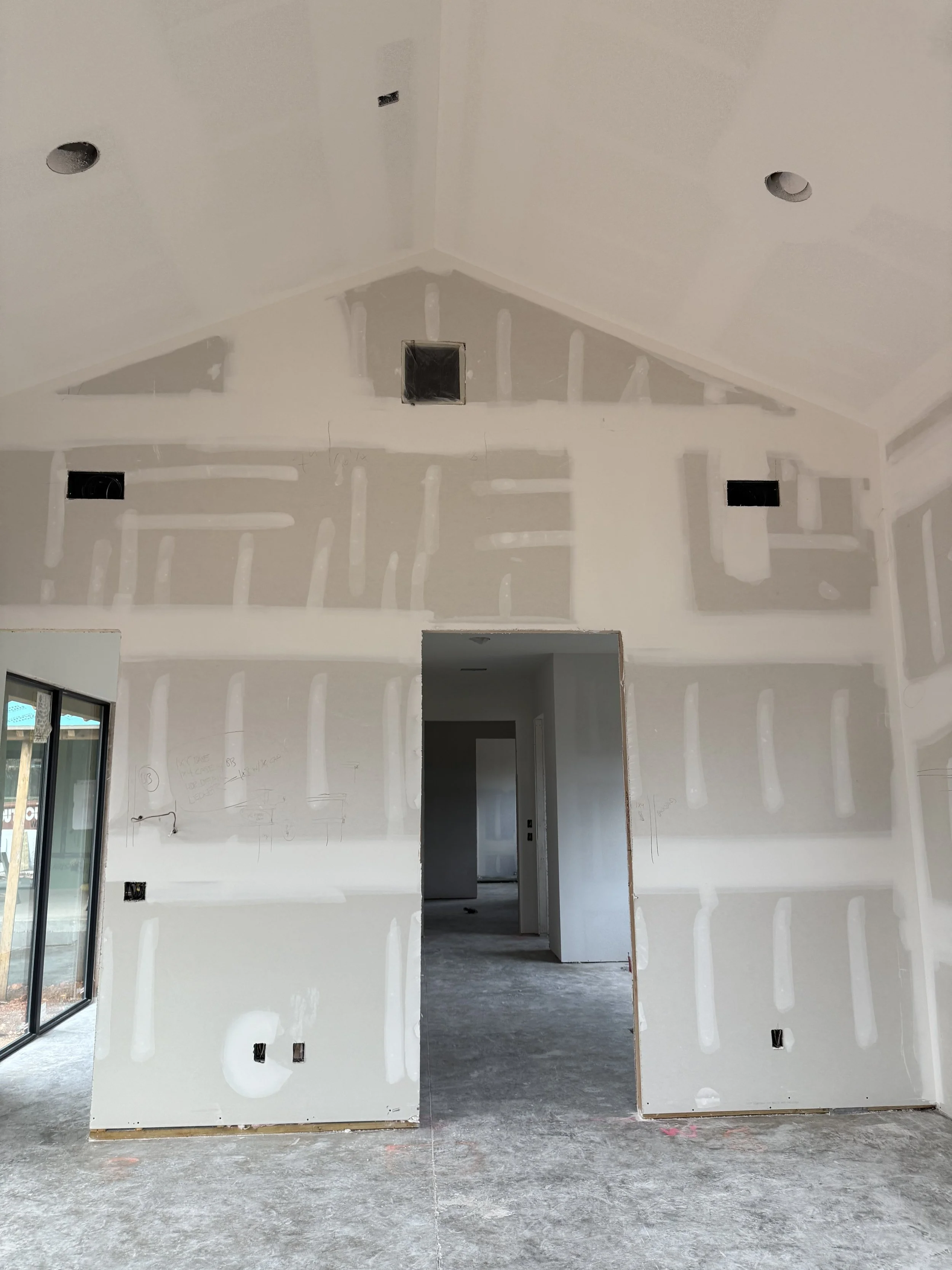 Interior of a house under construction with drywall walls and ceiling, visible electrical outlets, and unfinished flooring.