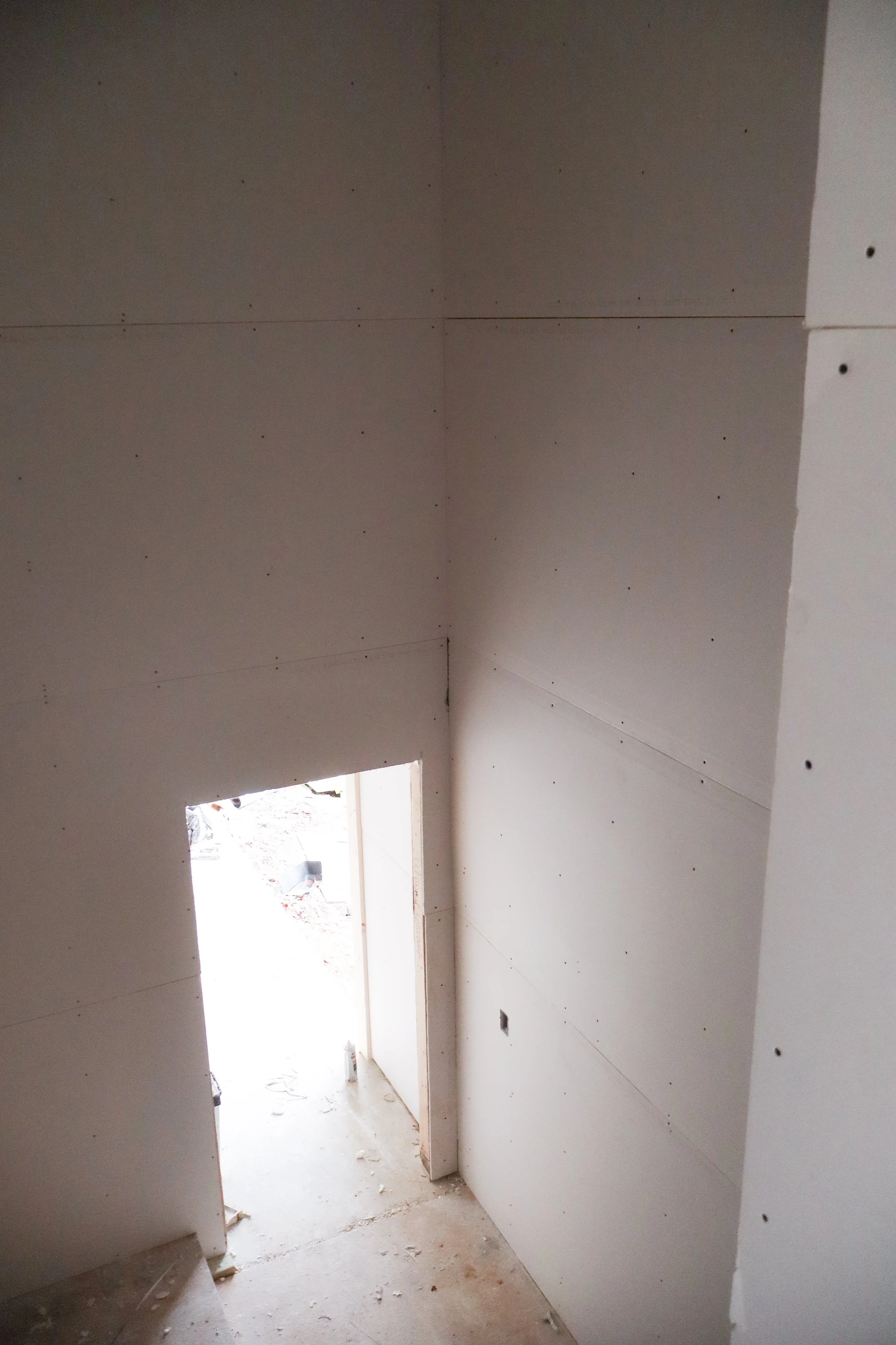 Interior view of a construction site with drywall being installed on walls and ceiling, with an open door leading outside.