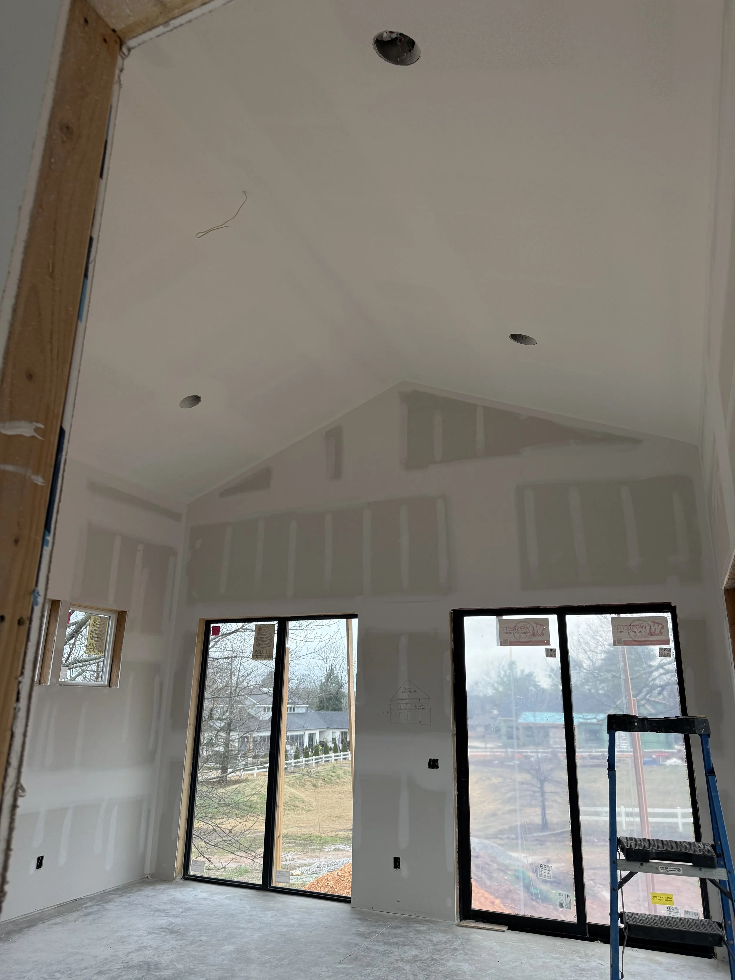Interior view of a room under construction with drywall installation, two large sliding glass doors, a small window, and a stepladder.