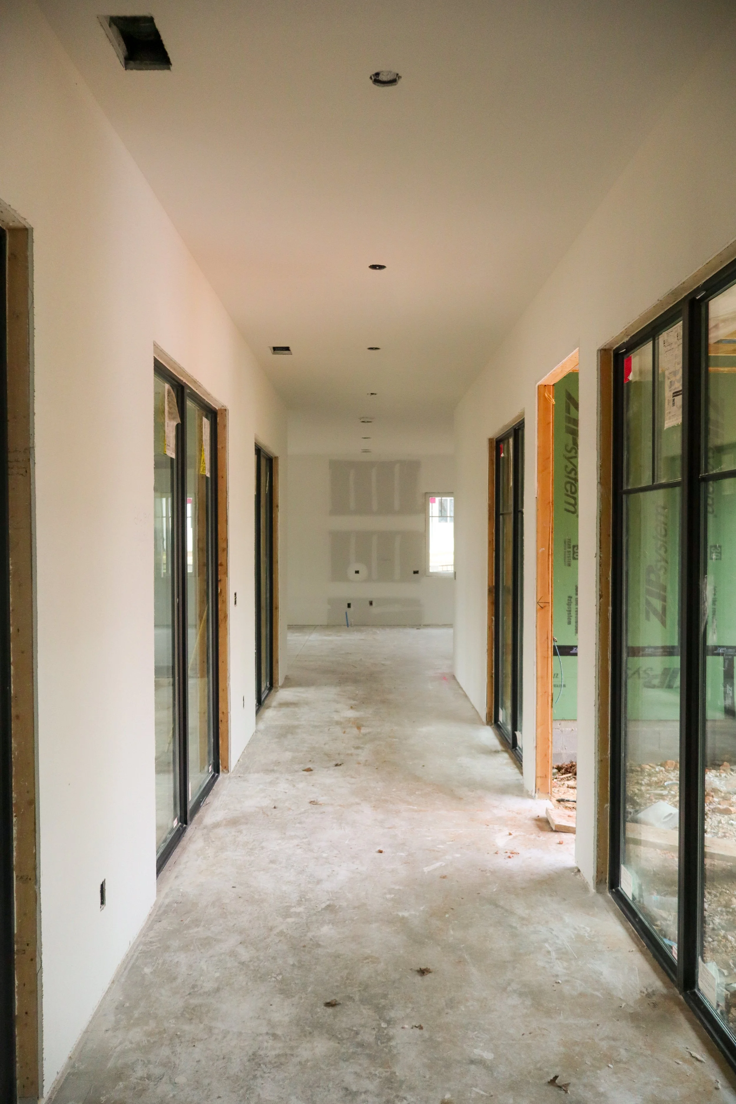 Interior of a house under construction, with a hallway, unfinished floor, and sliding glass doors on both sides leading to rooms, with some wall openings and visible construction materials.