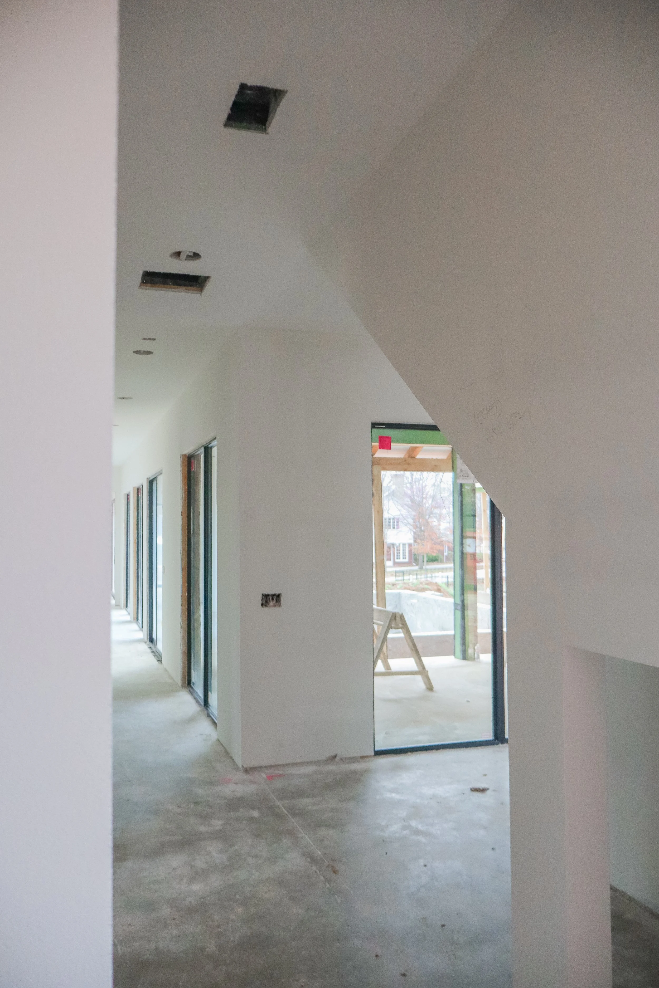 Interior of a house under construction, with white walls, unfinished concrete floor, and multiple doorways with black frames. The ceiling has several openings for light fixtures. Outside the window, houses and trees are visible.