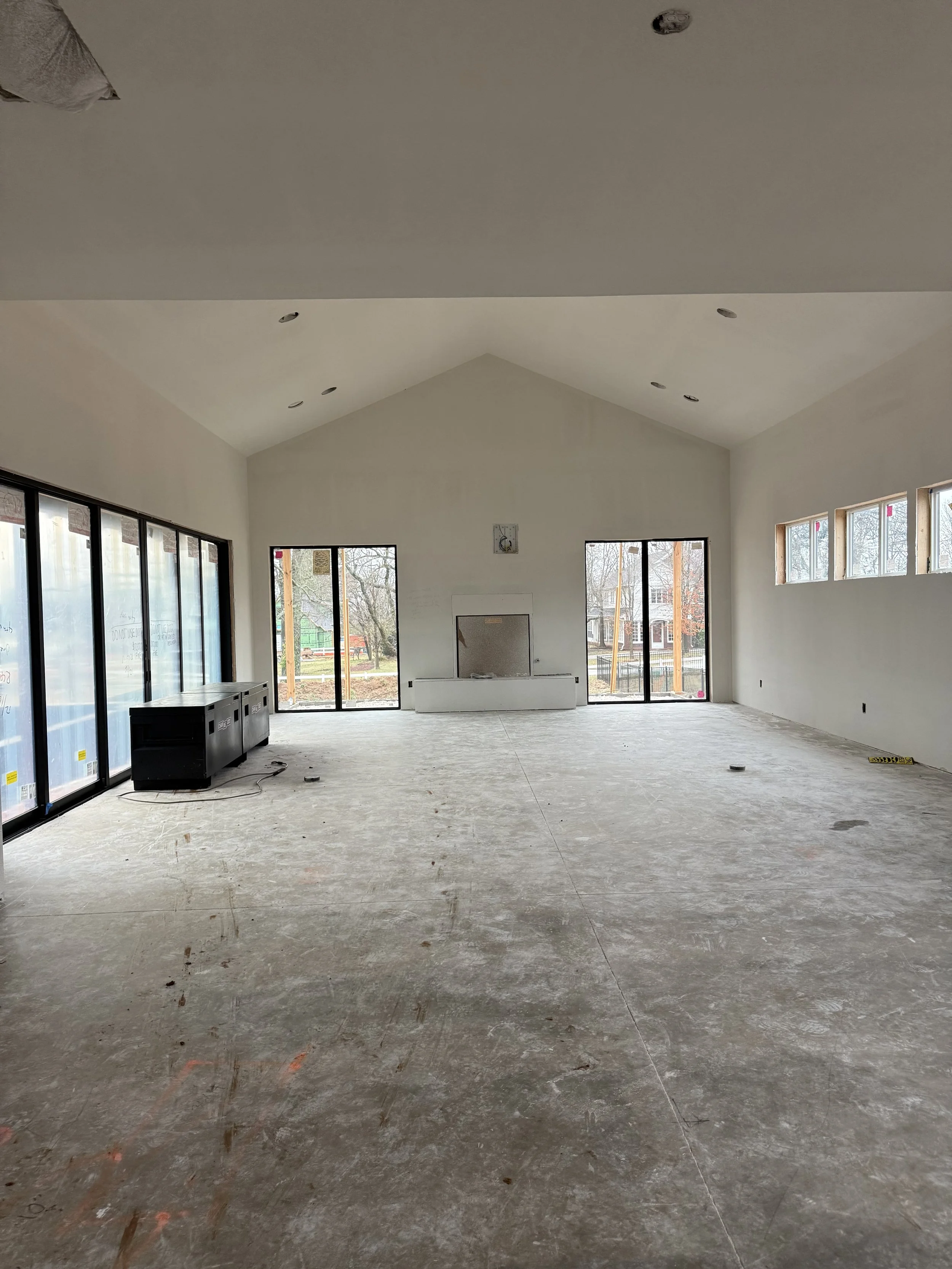 Interior of a room under construction with large windows, a vaulted ceiling, and a fireplace, with a bare concrete floor.