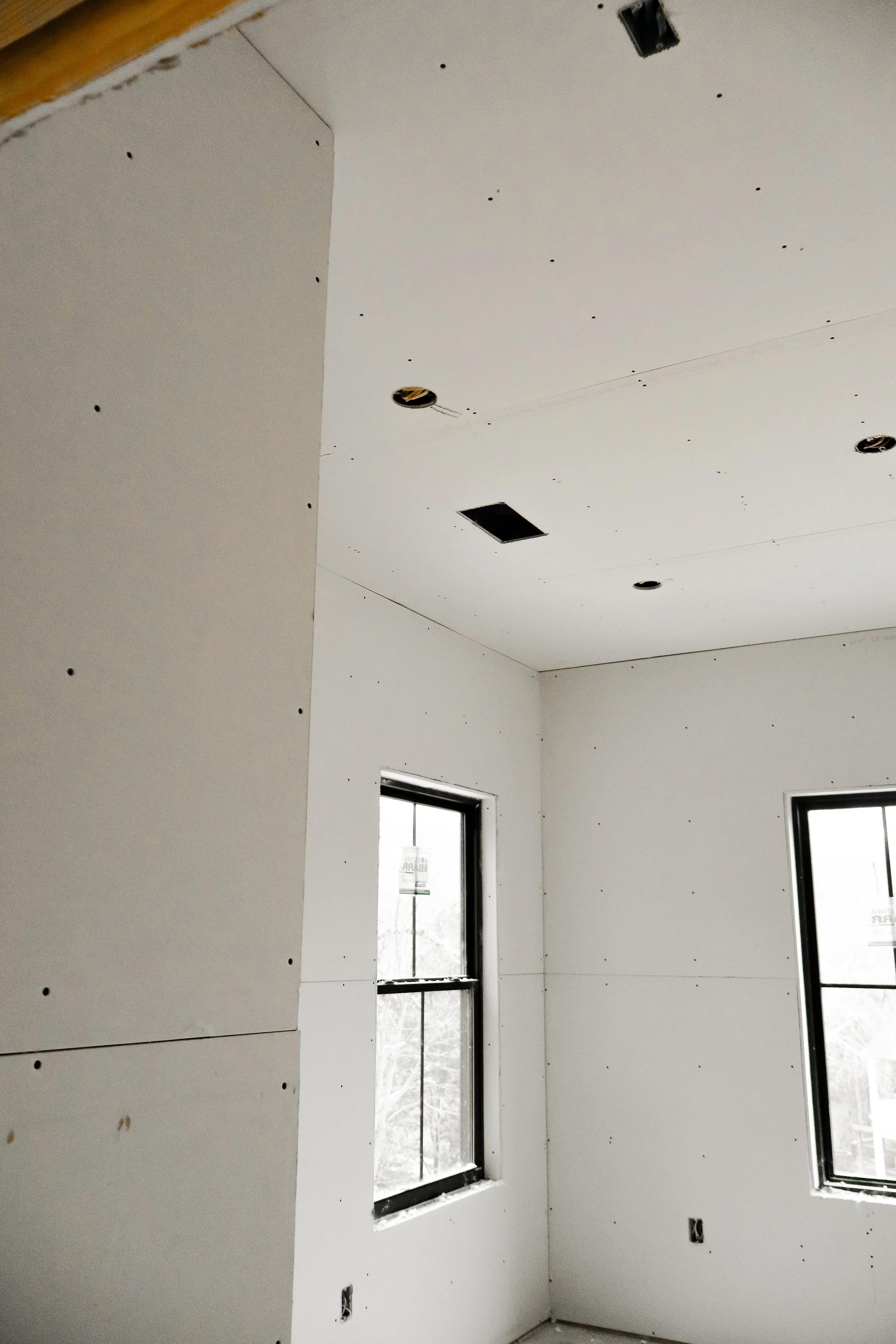Interior of a room under construction with white drywall installed, two windows, and ceiling openings for lights.