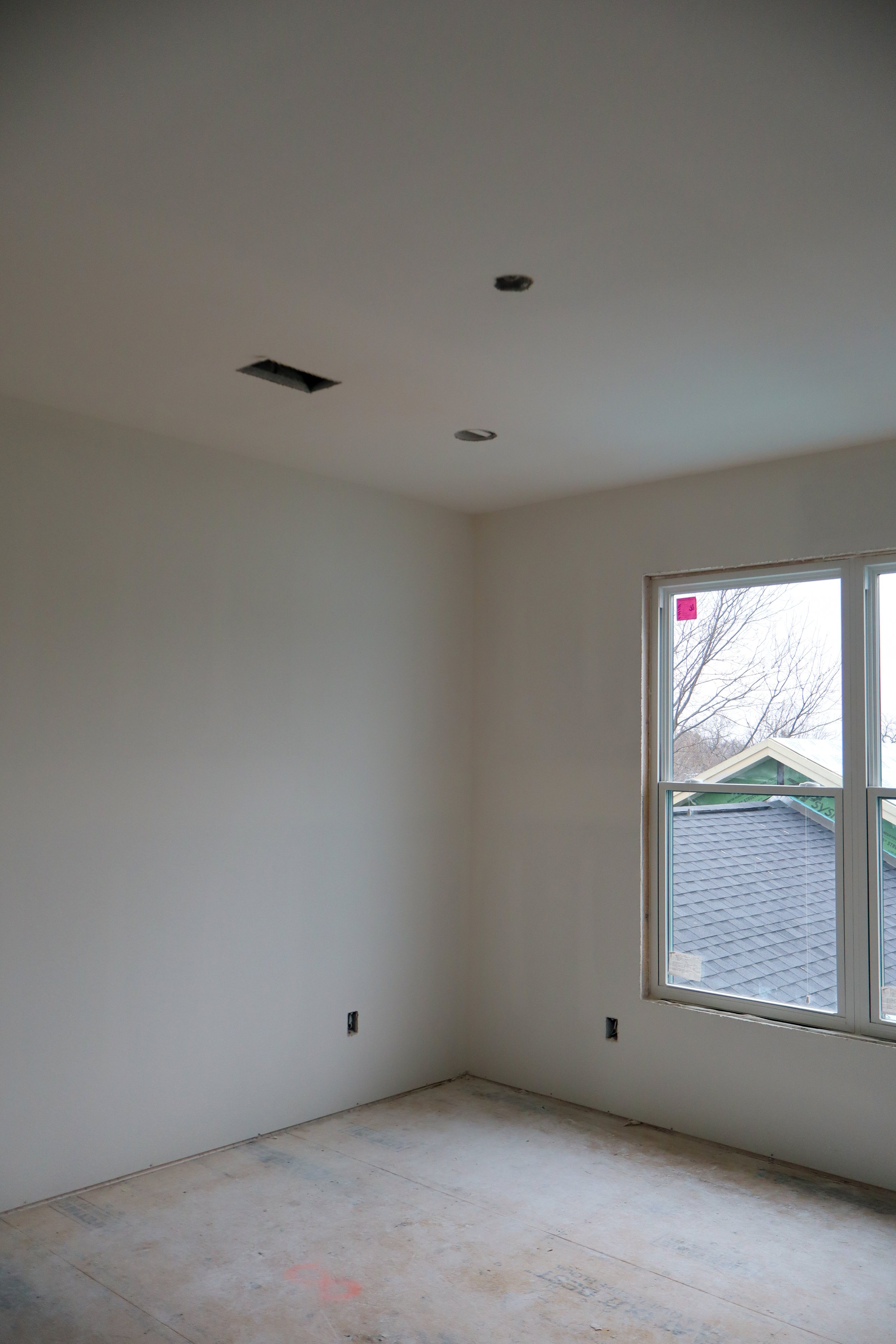 An empty room under construction with white walls, a large window showing a roof outside, and a ceiling with two open fixtures and one covered fixture.