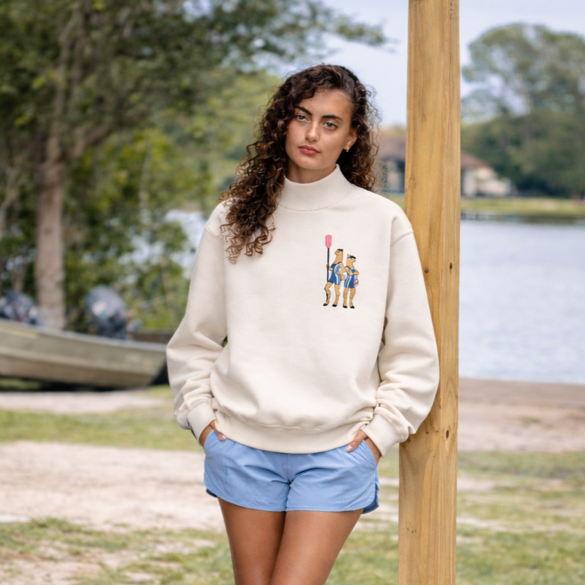 Young woman with curly brown hair standing outdoors near water and trees, wearing a cream sweatshirt with a mounted polo player graphic and light blue shorts.