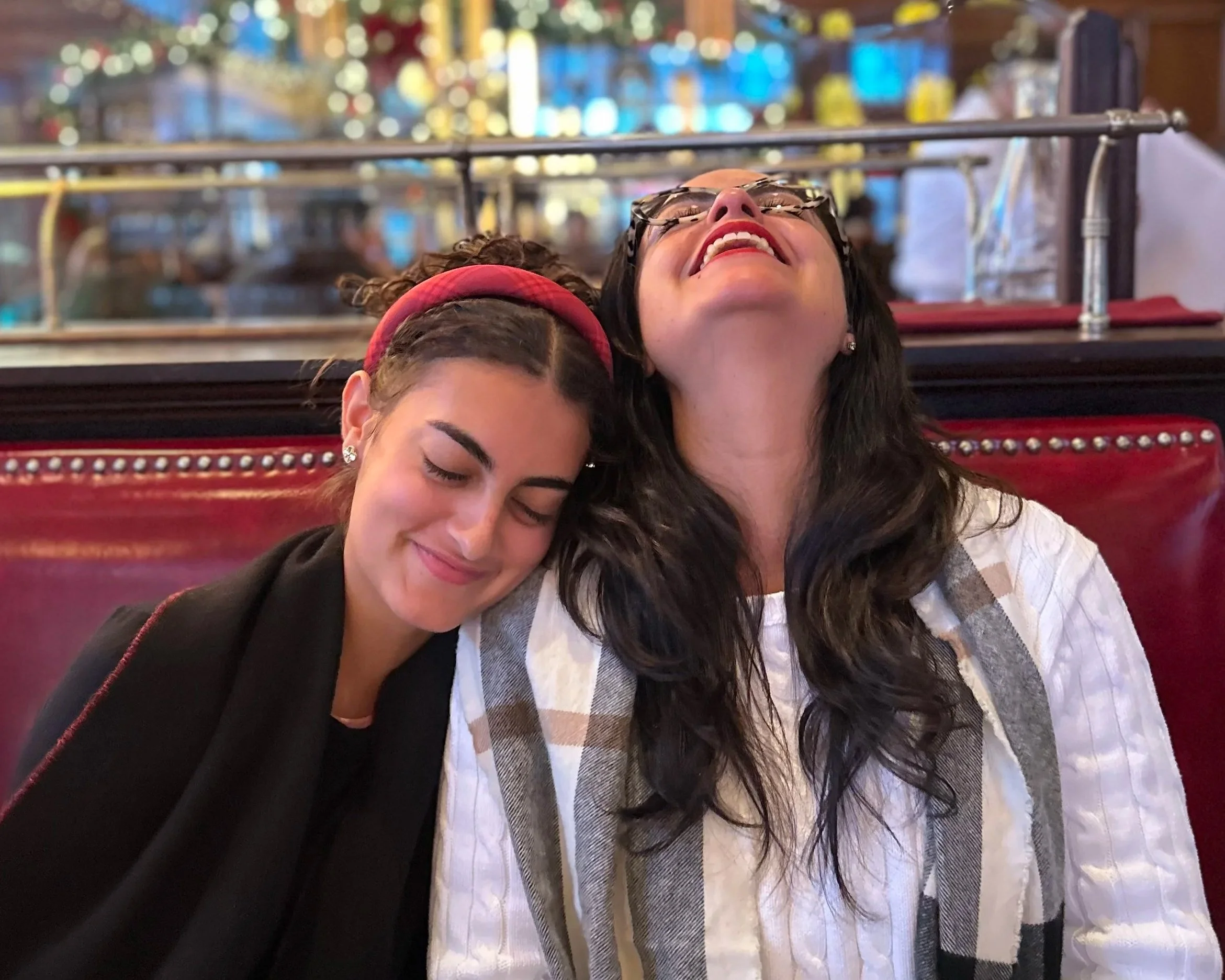 Two women sitting closely at a restaurant, smiling and enjoying each other's company with their eyes closed. The woman on the left has dark hair with a red headband, and the woman on the right has long dark hair and glasses.
