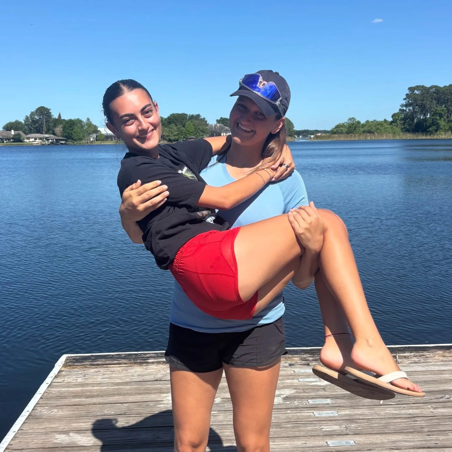 Two women on a dock by a lake, one holding the other in her arms; the woman being held has her arms around the other’s neck and is smiling, wearing red shorts, a black T-shirt, and flip-flops; the woman holding her is wearing a light blue T-shirt, black shorts, a dark cap with sunglasses, and is smiling at the camera.