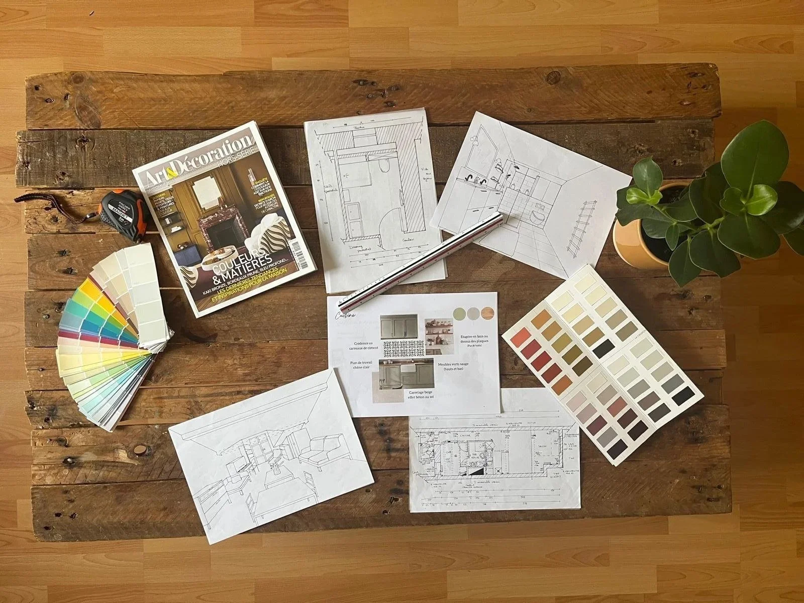 Une table en bois avec des plans d'architecture, une palette de couleurs, un magazine, un mètre ruban, des feuilles avec des croquis et des échantillons de couleurs, et une plante verte.