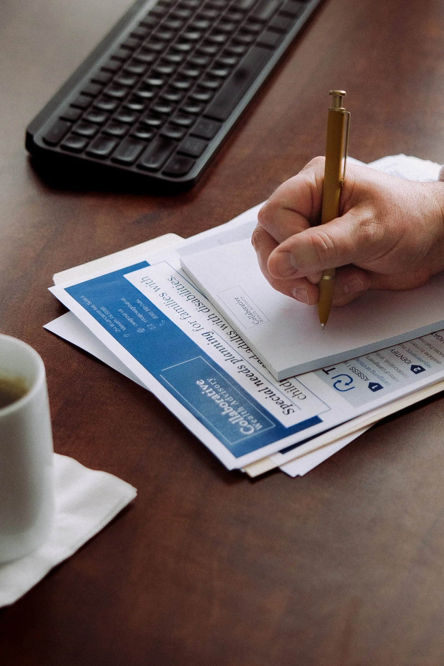 A person's hand writing on a notepad placed over some printed flyers on a wooden desk, with a computer keyboard and a cup of coffee nearby.