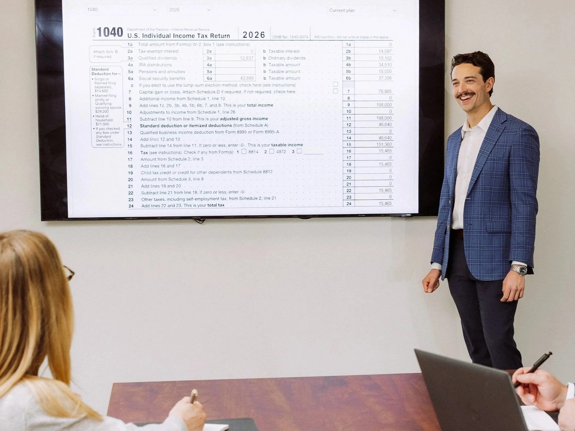 A man in a blue checkered blazer giving a presentation on a large screen showing a U.S. tax form. He is smiling, standing next to the screen, with two seated people in front of him, one with a laptop and the other taking notes.