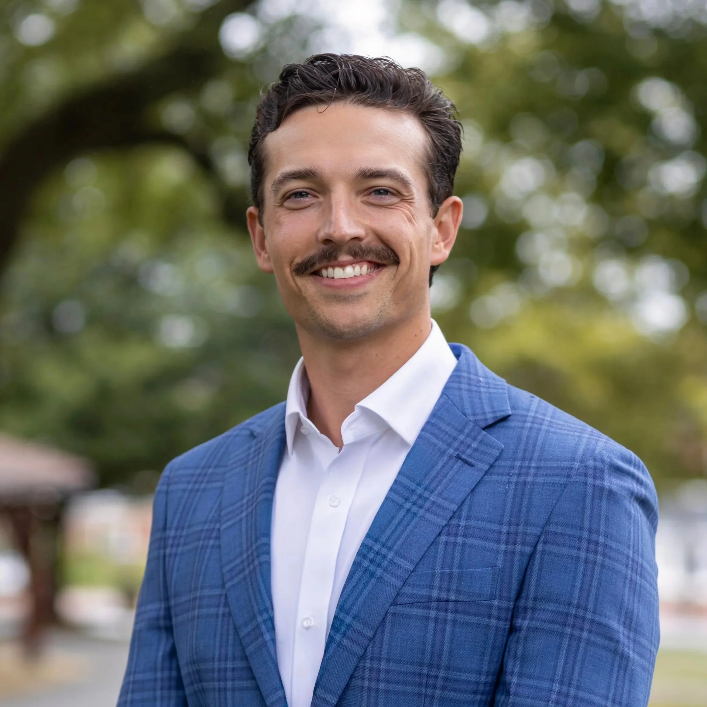 Smiling man with a mustache in a blue checked suit and white shirt outdoors with a blurred background of trees.