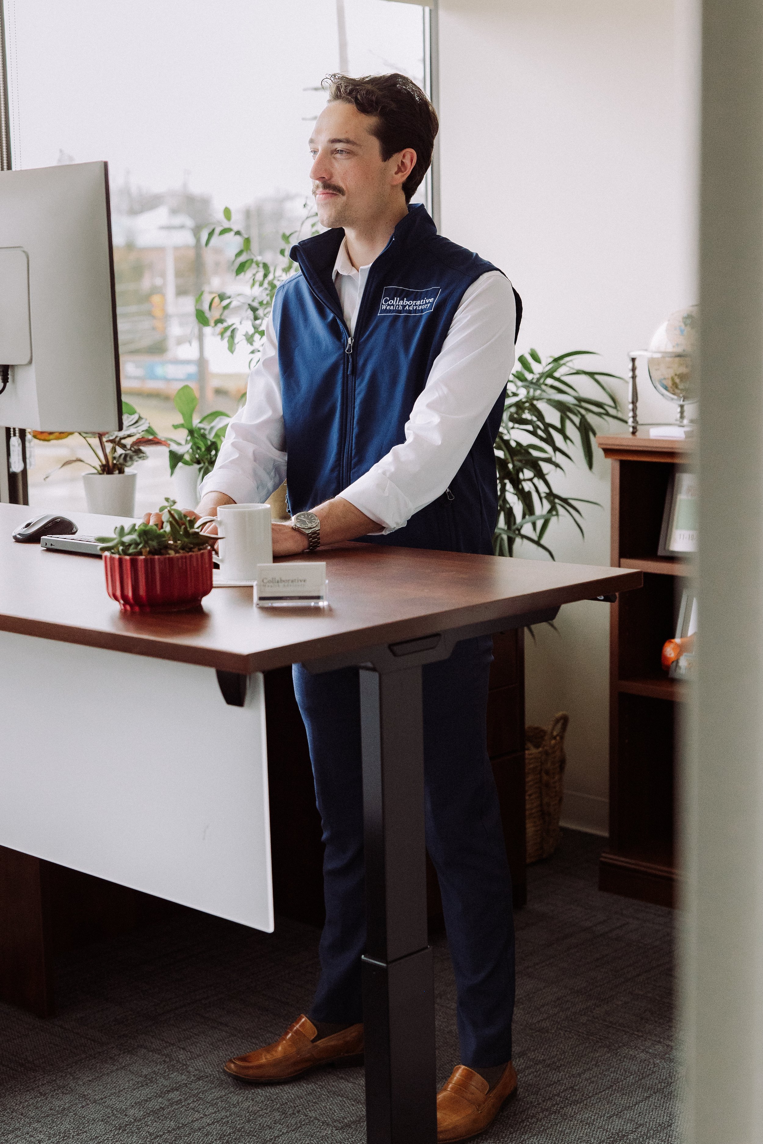 A man stands at a desk in an office, looking at a computer monitor, wearing a white shirt and a blue vest with a patch that reads 'Collaborative Wealth Advisors', with potted plants and office furniture in the background.