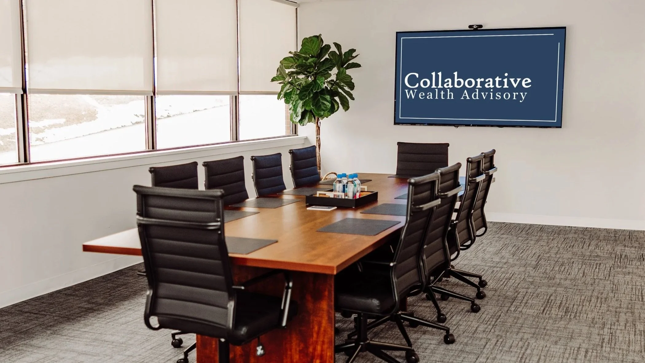 Large conference room with a wooden table, black office chairs, a large window with blinds letting in natural light, a potted plant, and a wall-mounted screen displaying 'Collaborative Wealth Advisory'.