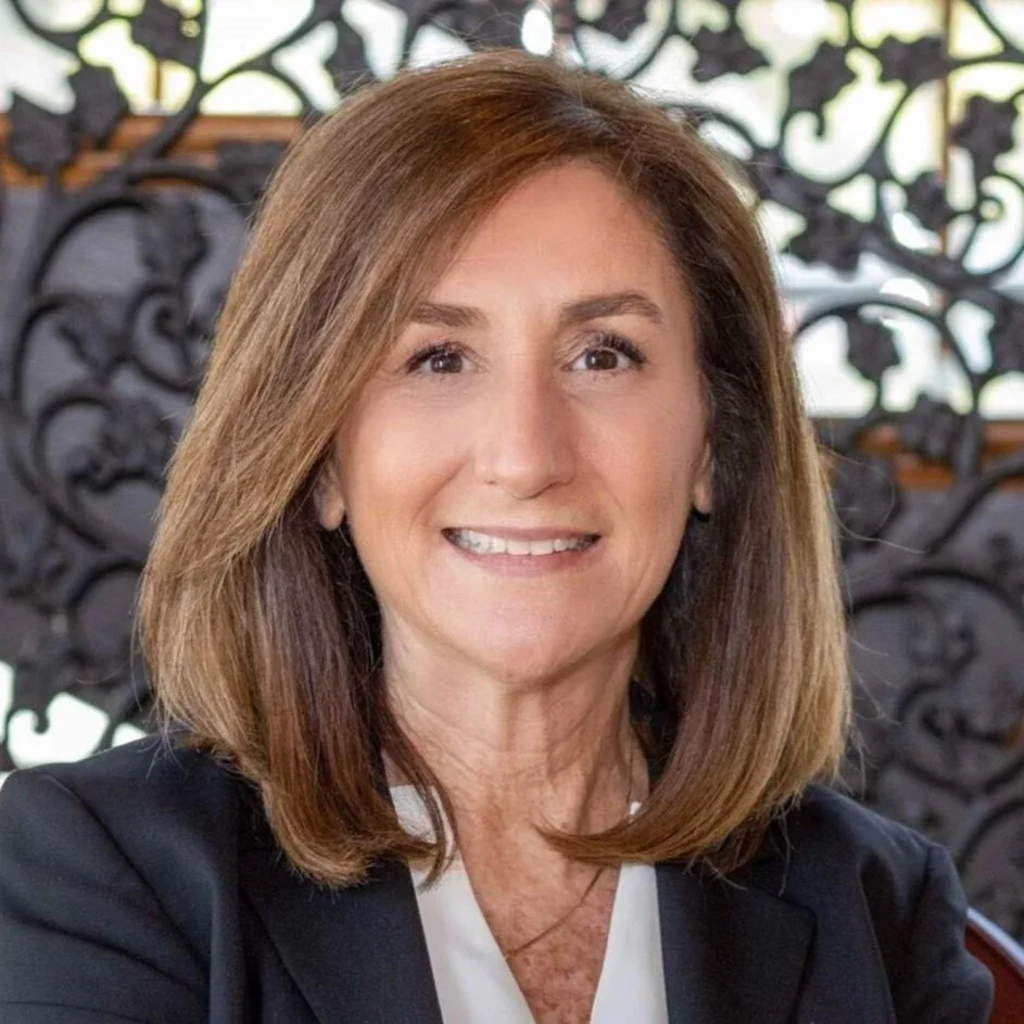 A woman with shoulder-length brown hair, smiling, wearing a black blazer and white blouse, with a decorative black wrought iron background.