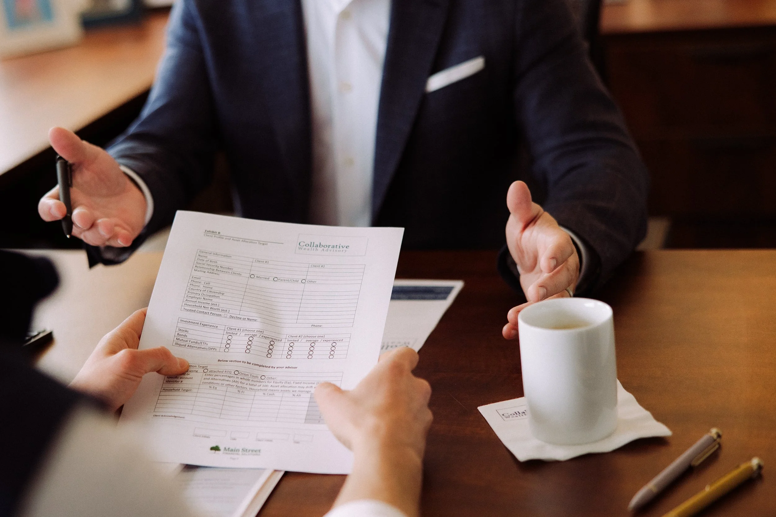 Two people sitting at a wooden table during a business meeting, one holding a financial document, with a white coffee mug and pens on the table.