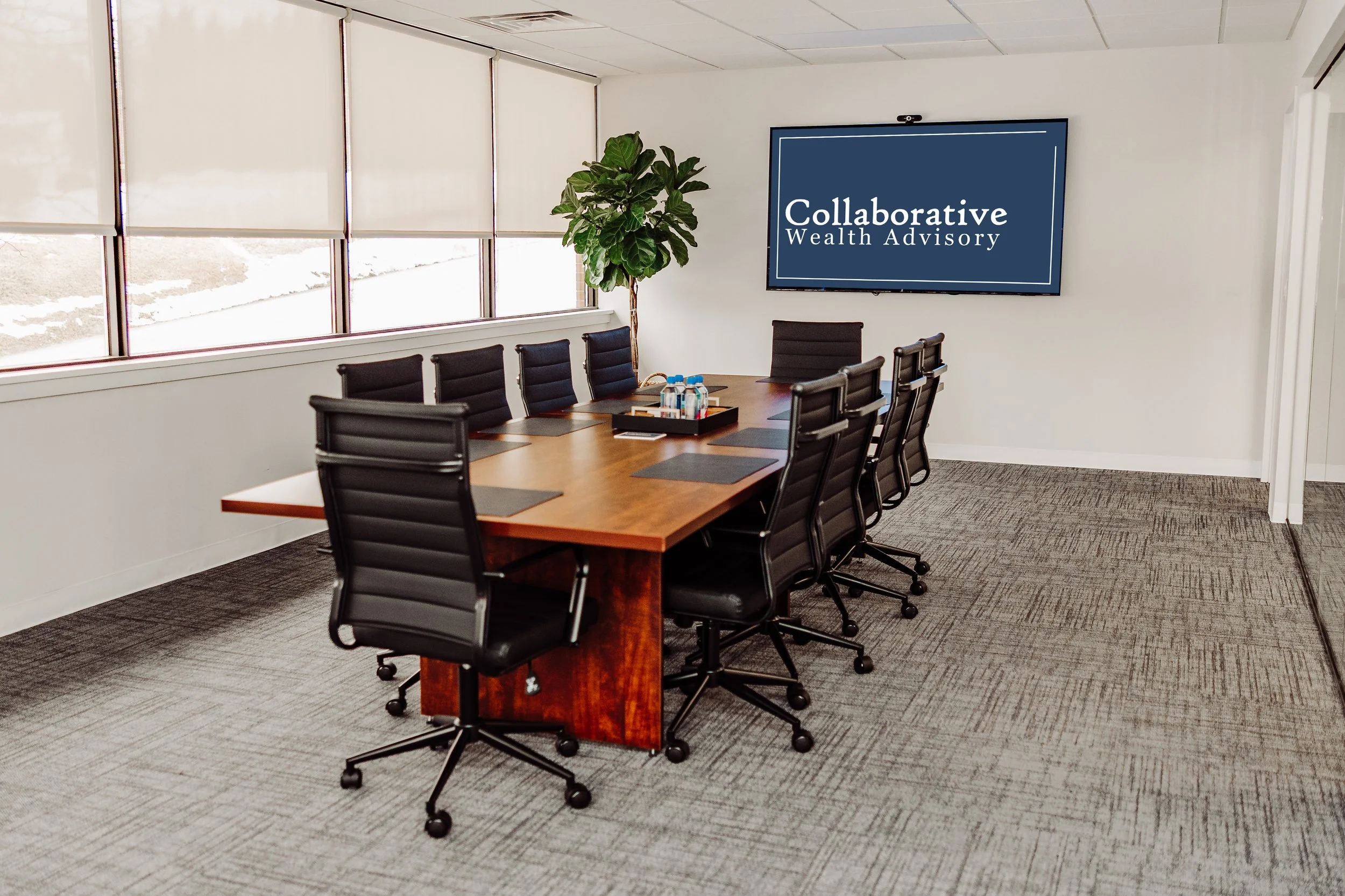 Empty conference room with a long wooden table, black office chairs, a potted plant, and a television screen displaying 'Collaborative Wealth Advisory'.