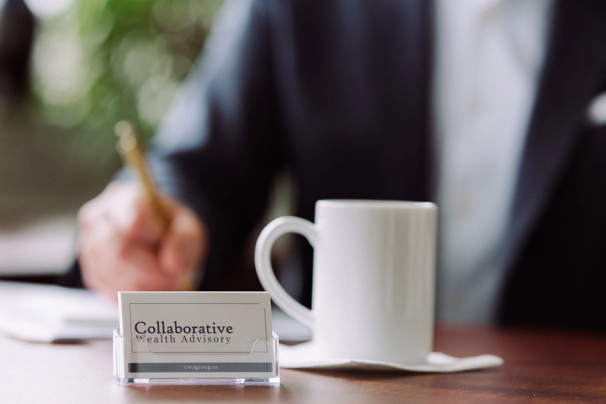 Close-up of a business card labeled 'Collaborative Wealth Advisory' on a table, with a white coffee mug in the background and a person in a dark suit writing with a gold pen.