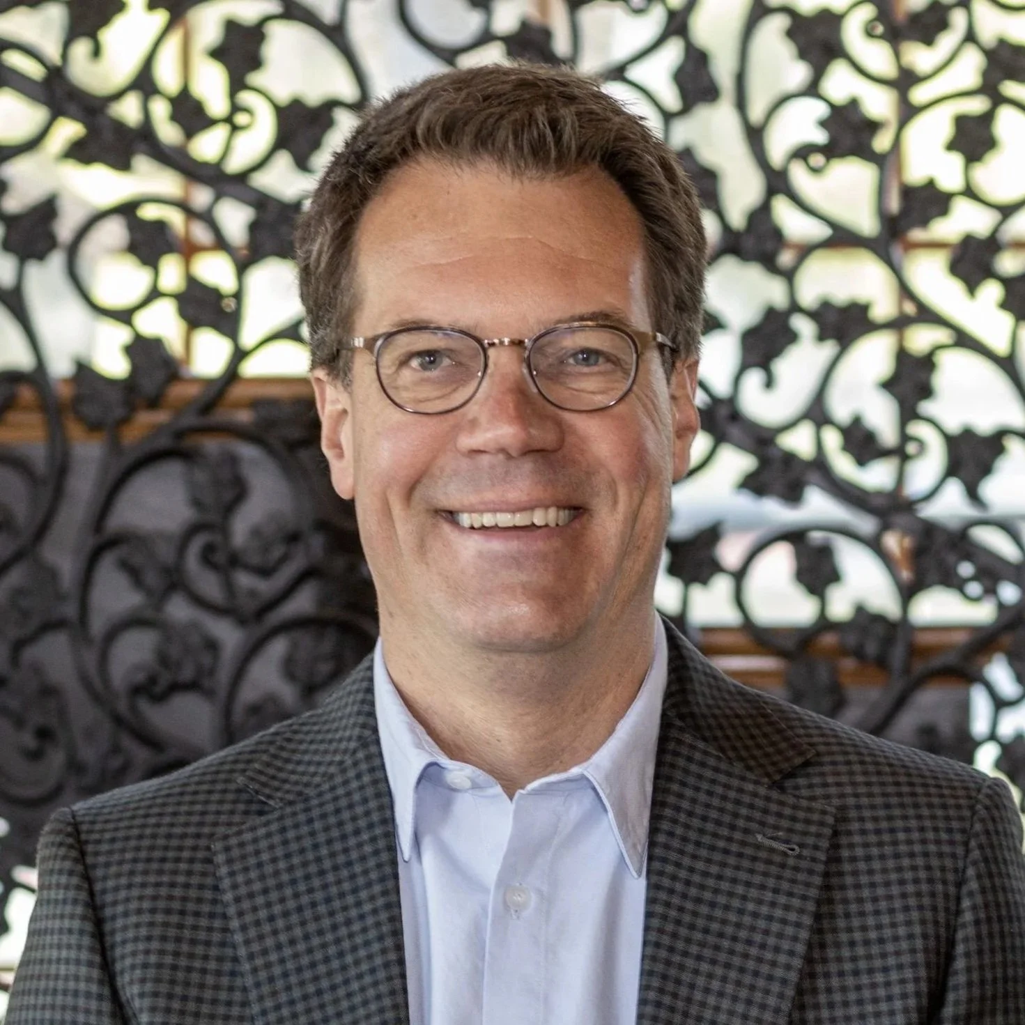 A smiling man with glasses and brown hair, wearing a checkered blazer and white shirt, standing in front of an ornate black wrought iron background.