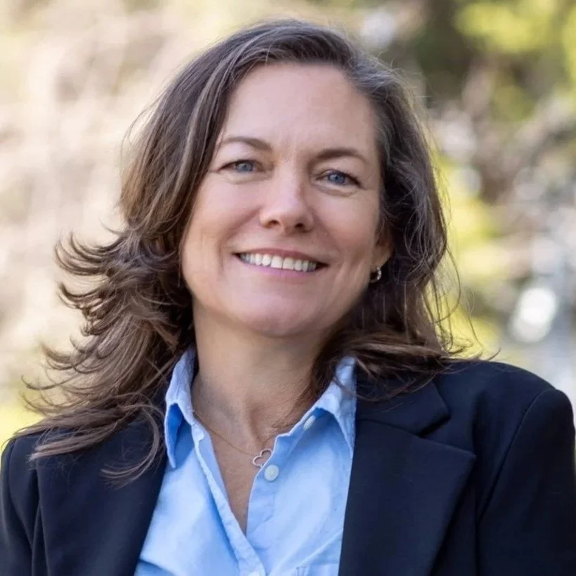 A smiling woman with shoulder-length brown hair, wearing a black blazer over a light blue collared shirt, standing outdoors with blurred trees in the background.