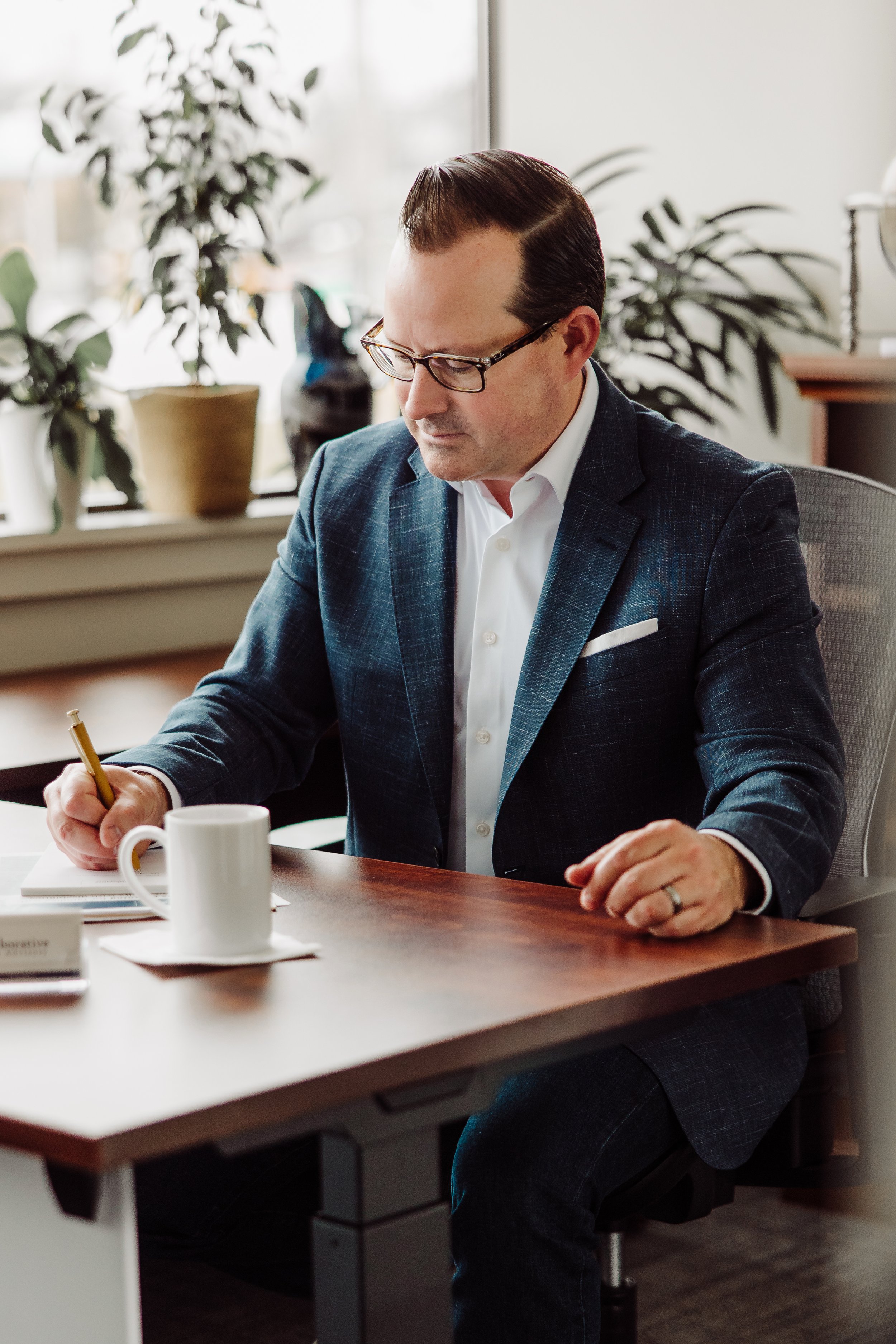 A man in a business suit and glasses sitting at a wooden desk, writing in a notebook with a gold pen, with a coffee mug nearby, in an office with plants and a window in the background.