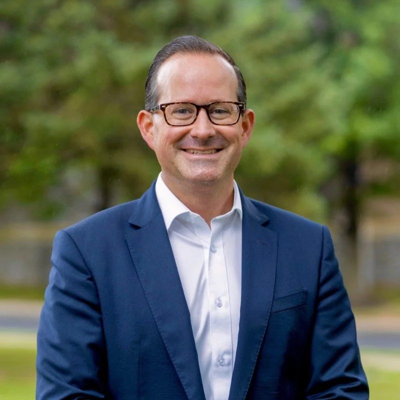 A man in a navy blazer and white shirt, wearing glasses, smiling outdoors with blurred green trees in the background.