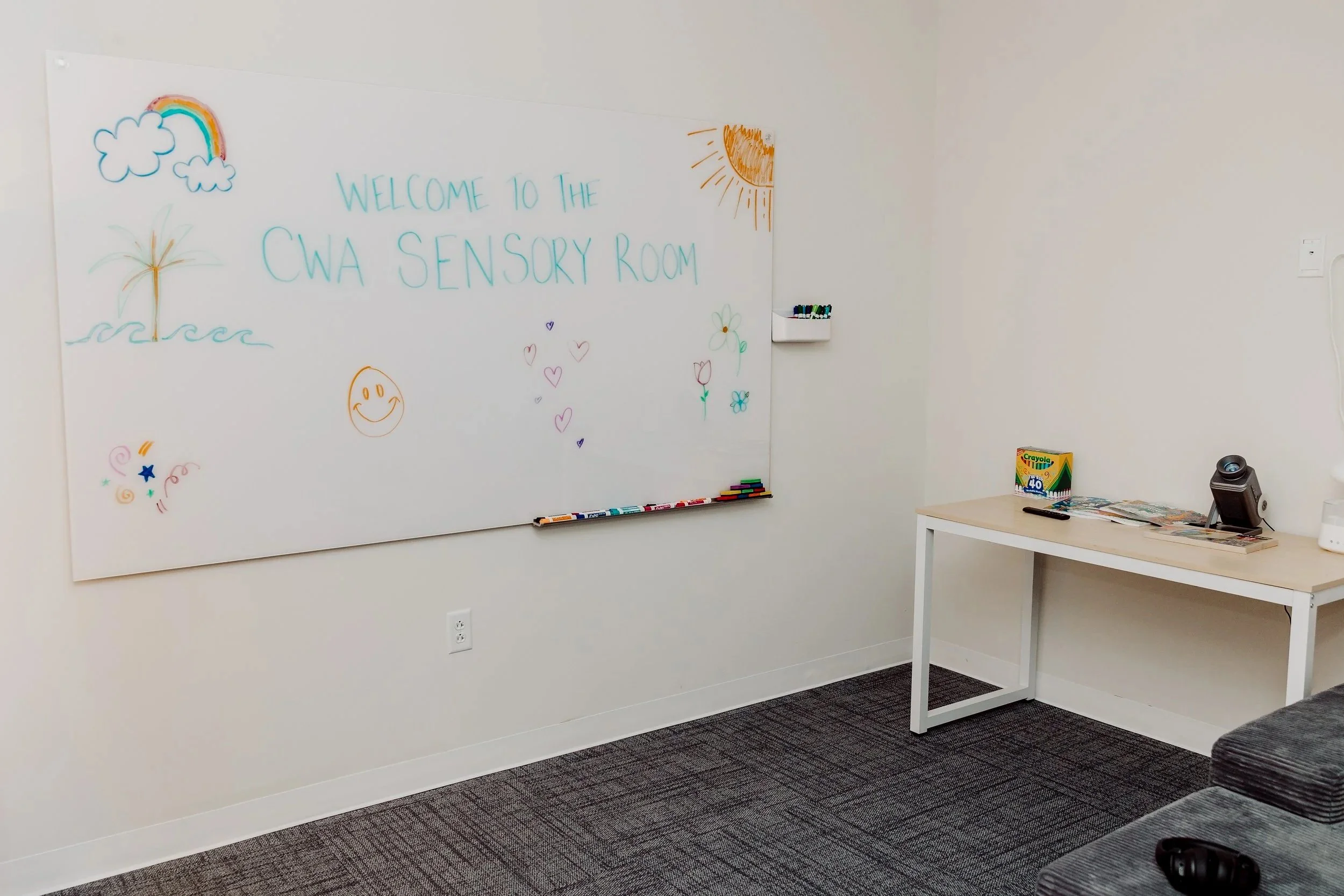 Whiteboard with colorful drawings of a rainbow, sun, boat, flowers, hearts, and a smiley face, with the handwritten message 'Welcome to the CWA Sensory Room' in blue. A small white shelf holding markers is attached to the whiteboard. To the right, a white table holds children's coloring supplies, a box of Crayola crayons, a remote control, some magazines, and a small speaker. The room has gray carpet and off-white walls.
