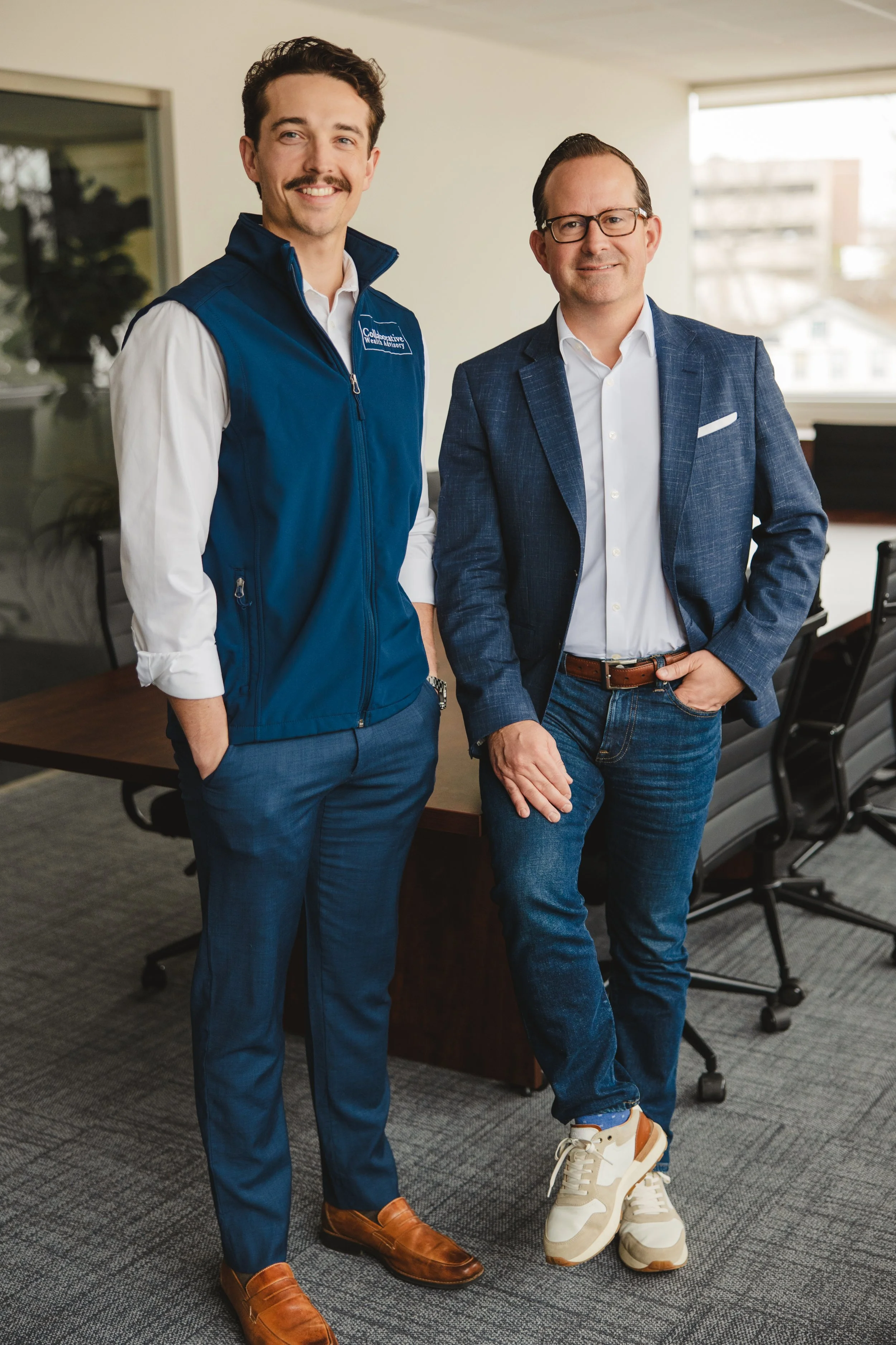 Two men standing in an office, smiling at the camera. One wears a white shirt, blue vest, and blue pants, while the other wears a white shirt, blue blazer, and jeans with sneakers.