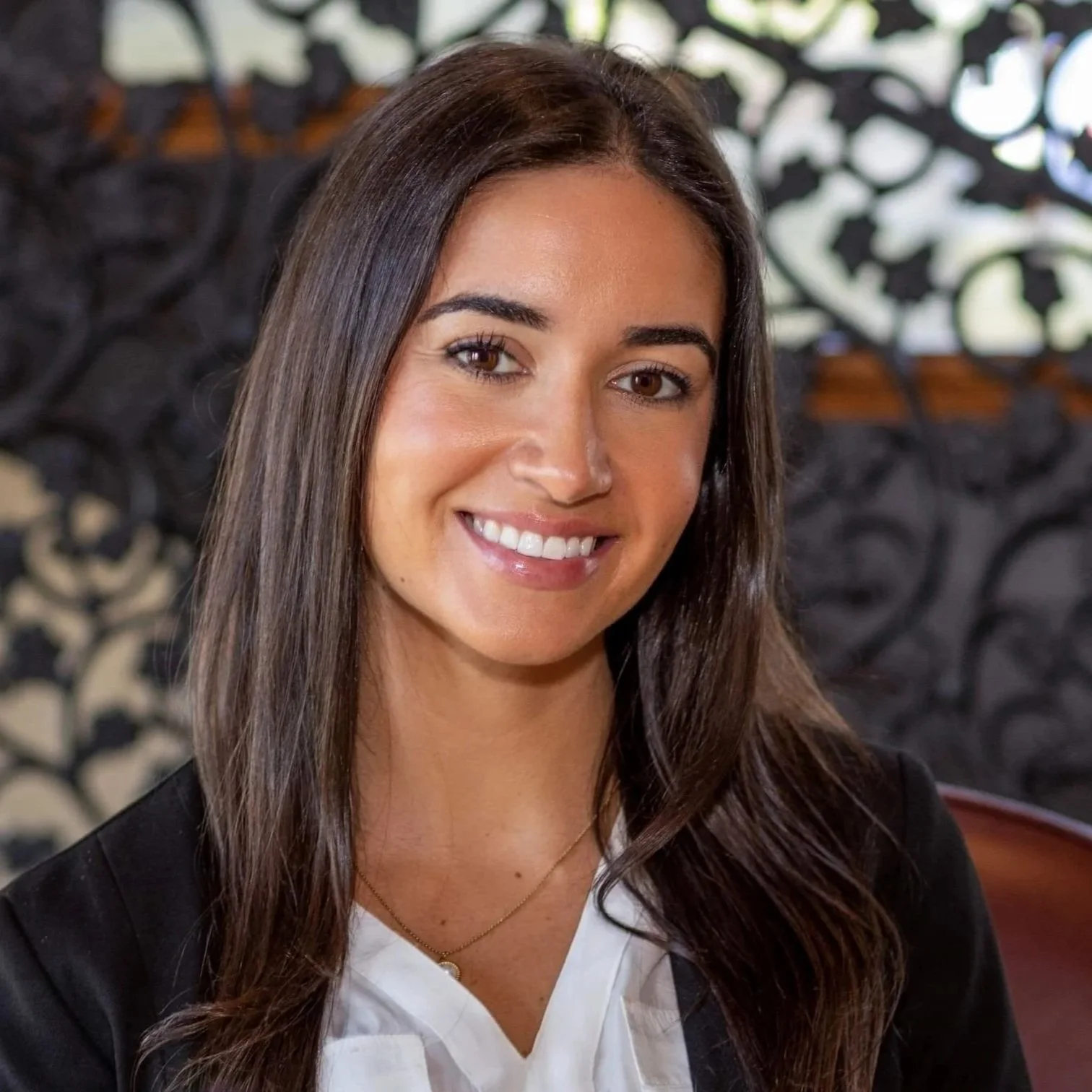 A smiling woman with long brown hair, wearing a white blouse and black blazer, sitting in front of an ornate black screen.