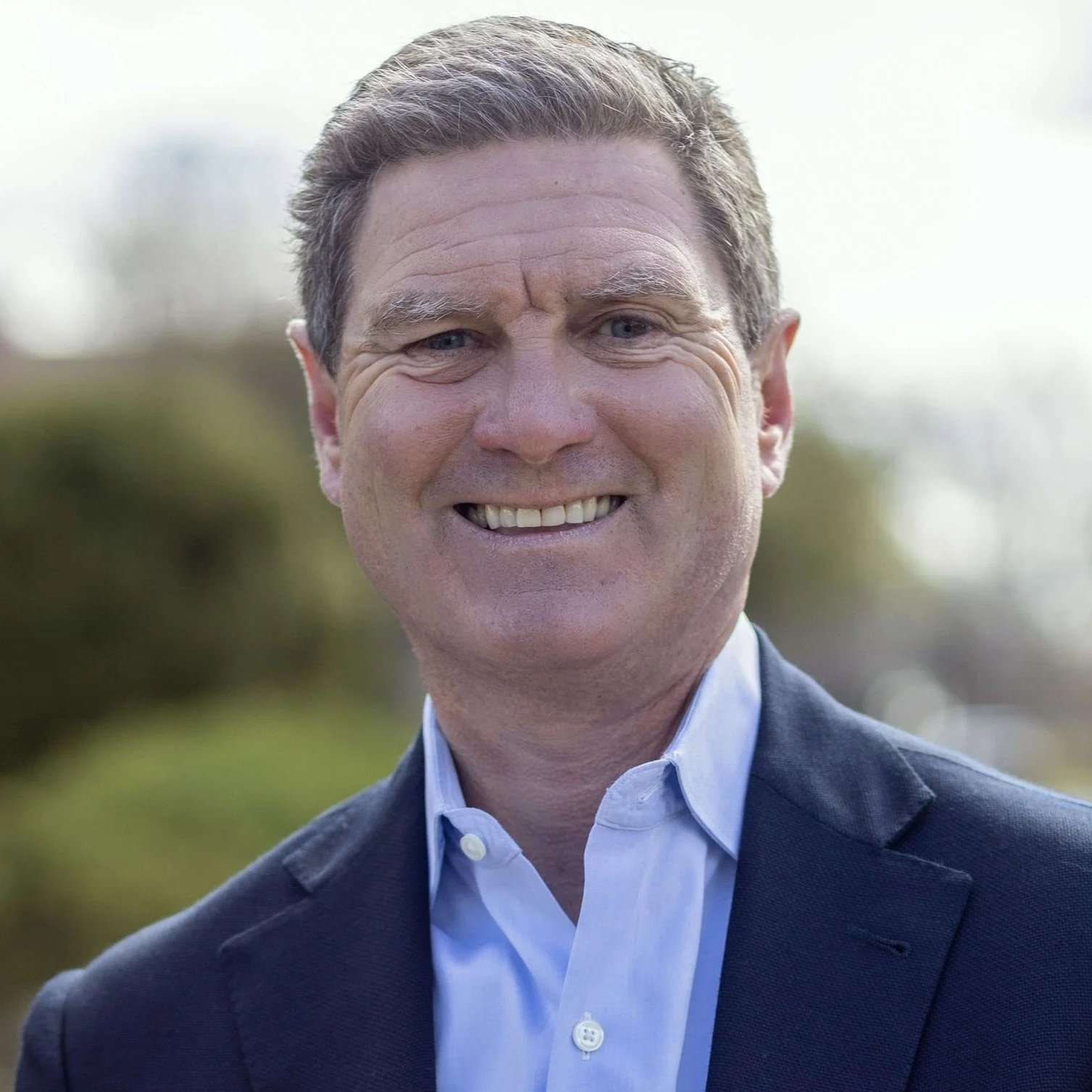 Headshot of a smiling man in a suit outdoors with a blurred background.