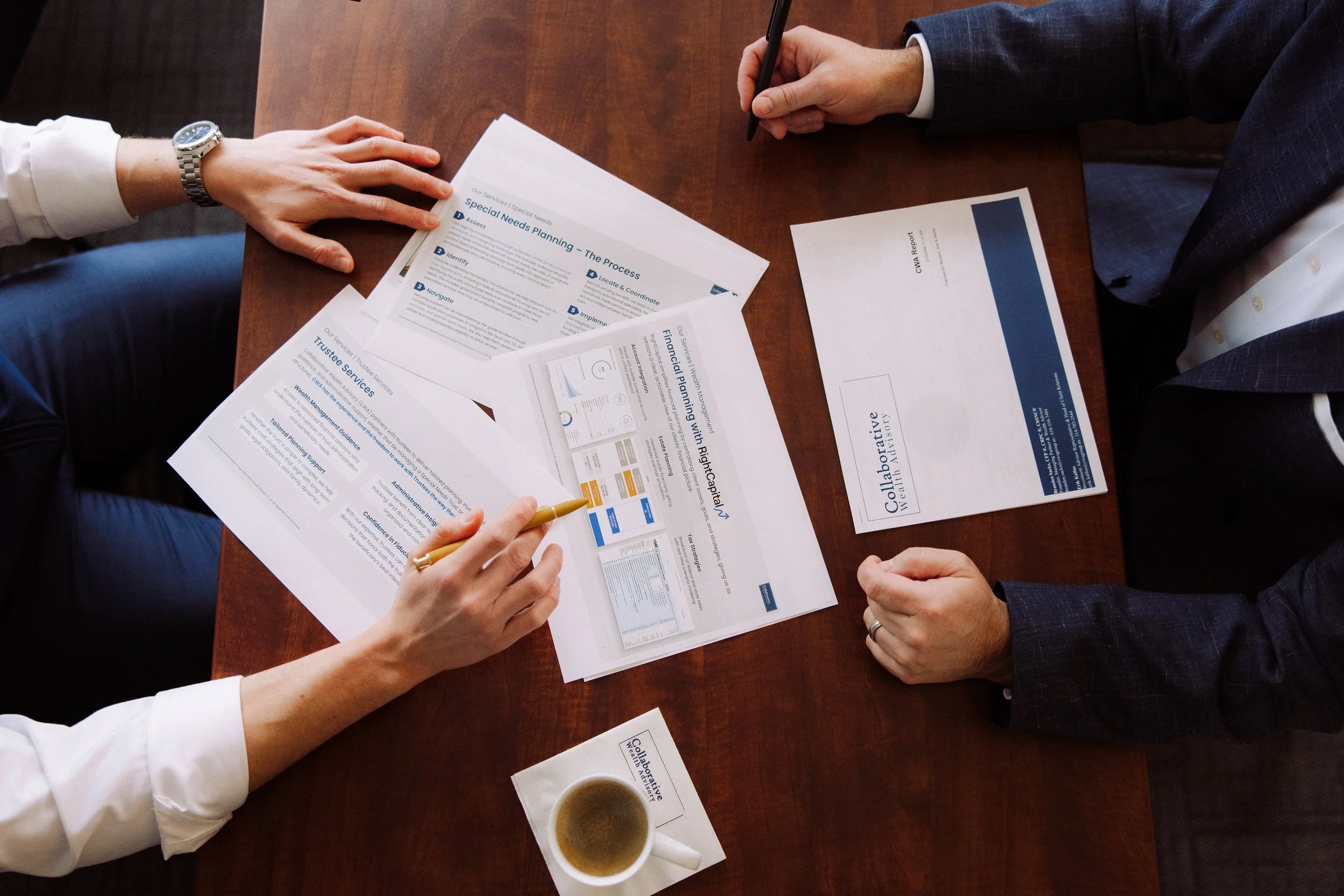 Two people in business attire sitting at a wooden table, reviewing and discussing printed documents and reports on planning and services, with a cup of coffee nearby.