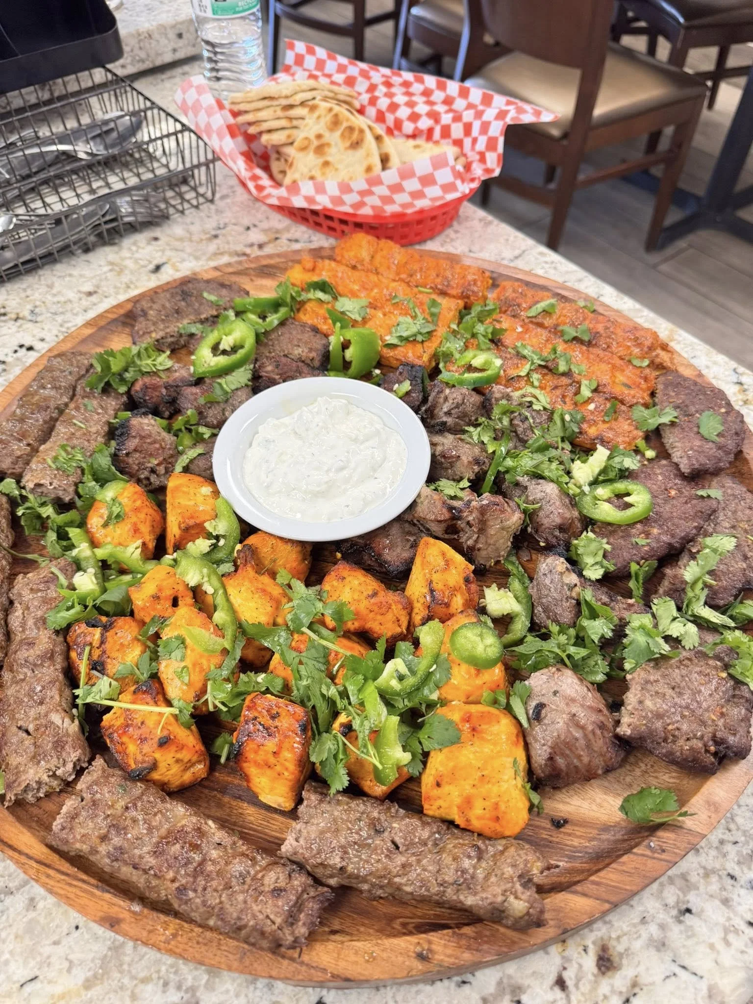 Large round wooden platter with grilled meats, roasted sweet potatoes, and green peppers, garnished with cilantro and a small bowl of white sauce. In the background, a basket with grilled flatbread, a water bottle, and a table with chairs.